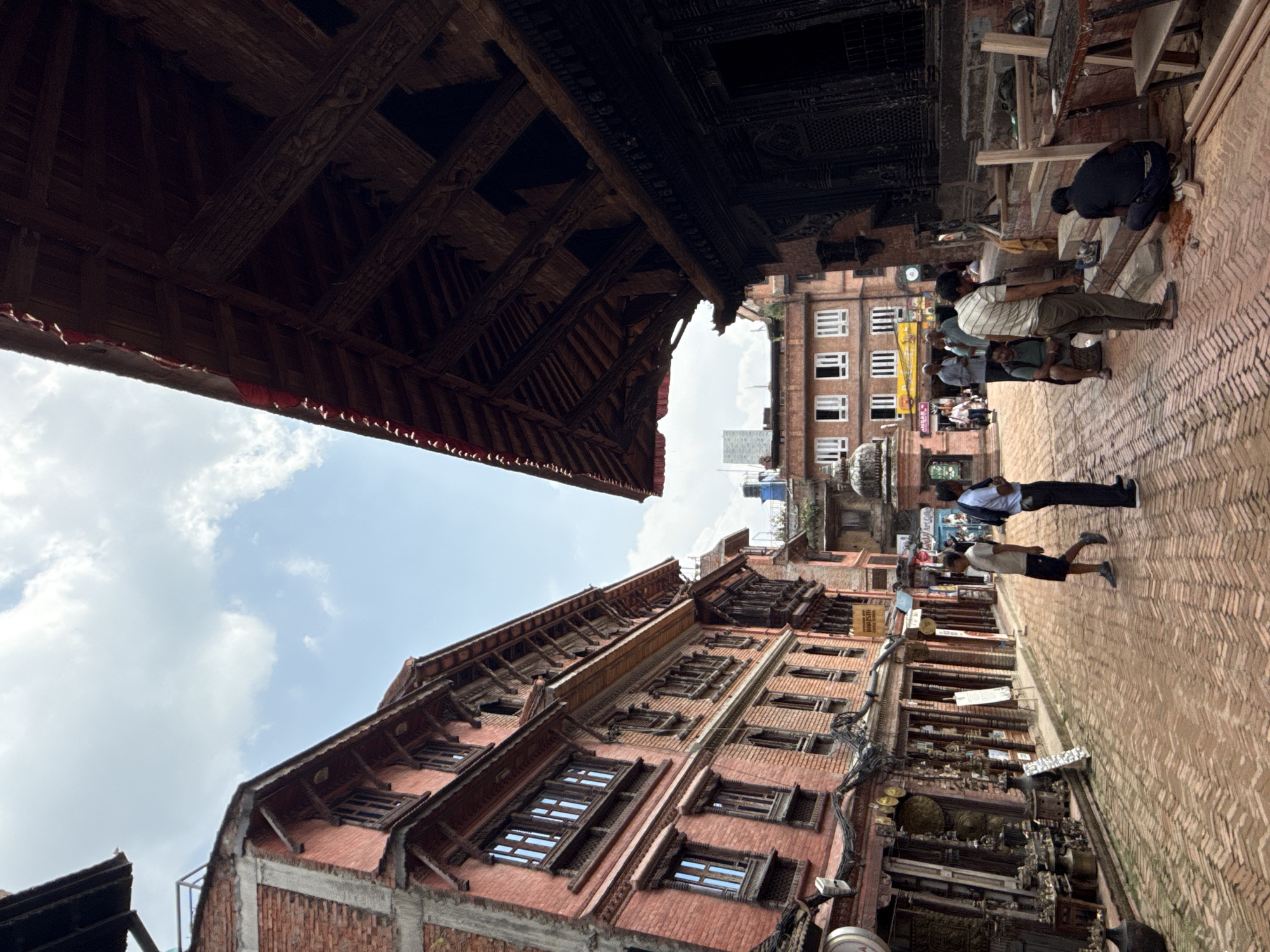The image shows a street scene in Bhaktapur, Nepal. The camera is angled upwards, capturing the architectural details of traditional brick buildings with ornate wooden window frames and overhanging structures. The sky with scattered clouds is visible between the buildings. On the right side of the image, people are walking along a brick-paved street. Some individuals are engaged in construction work.