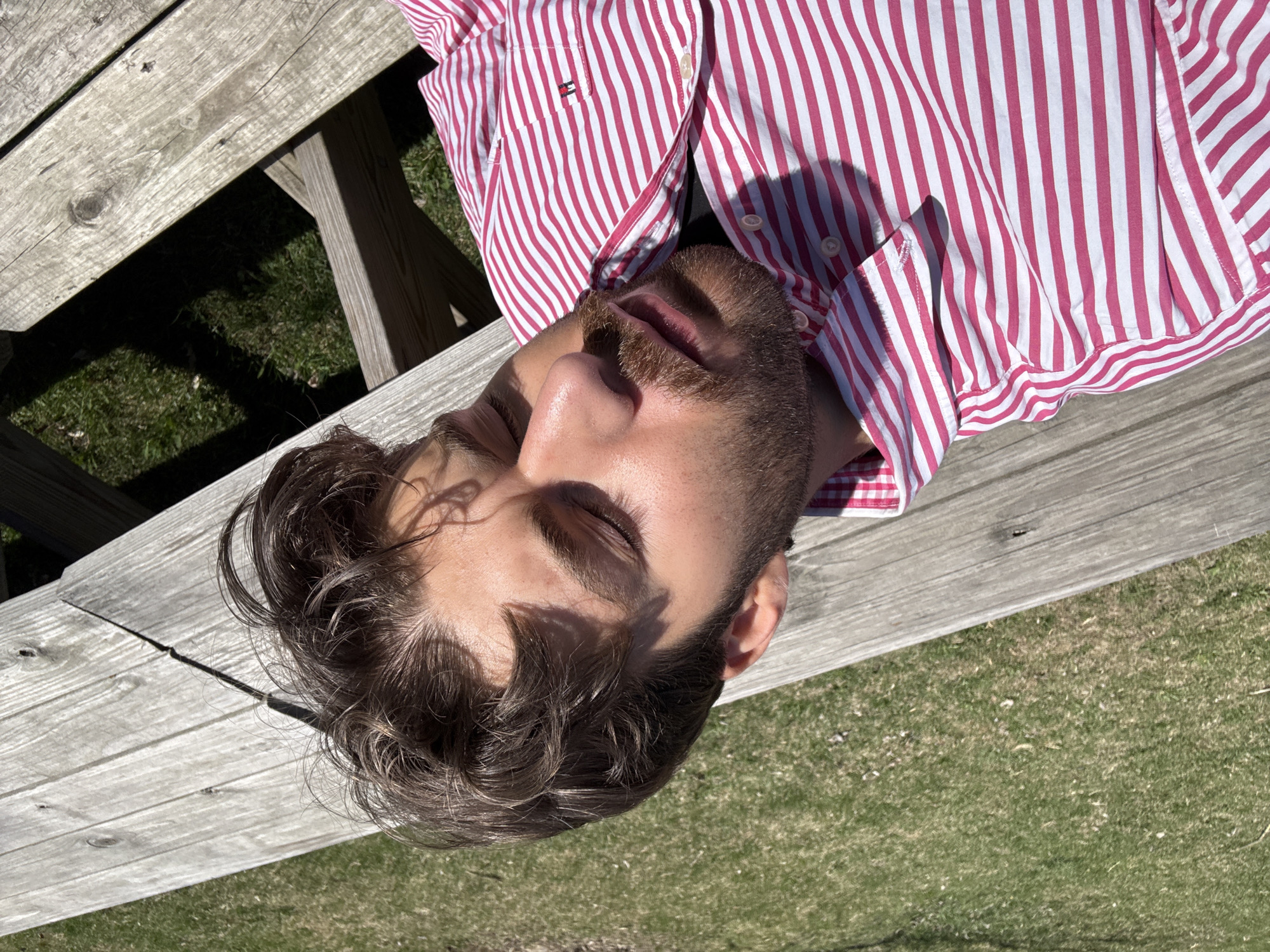 An overhead shot shows a man resting or sleeping on a weathered wooden picnic table in an outdoor setting. He is positioned diagonally across the frame, with his head towards the bottom-left and body extending to the top-right. The man has closed eyes, short brown hair, and a neatly trimmed beard. He is wearing a red and white striped long-sleeved button-up shirt. A small Tommy Hilfiger logo is visible on the shirt's left chest area. Bright sunlight illuminates his face and shirt, casting distinct shadows, indicating a clear, sunny day. Green grass is visible on either side of the picnic table, suggesting a park or garden environment. The overall mood is one of peace and relaxation. The location context provided is Montreal, Canada.