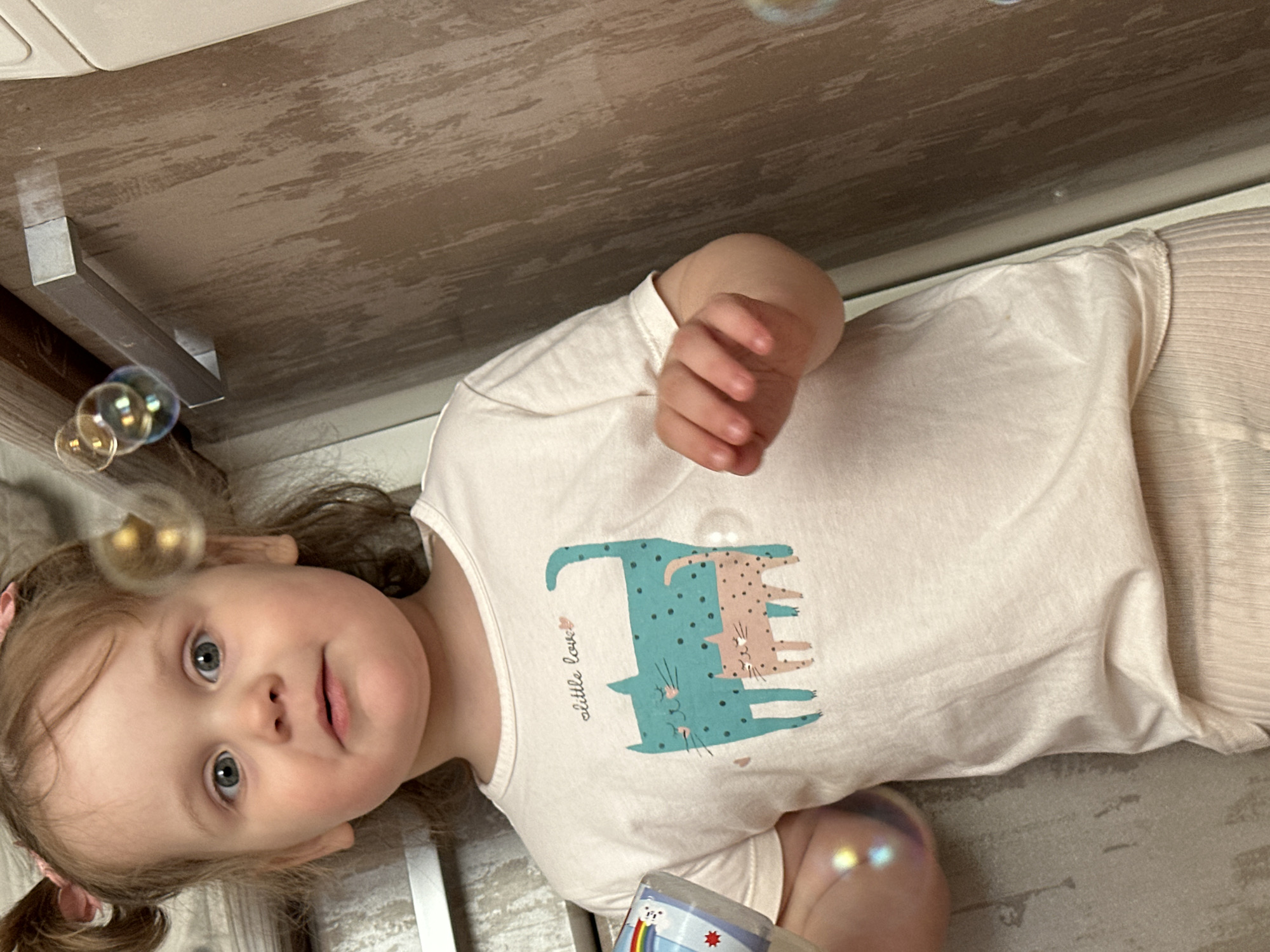 A young child with pigtails lies on their back indoors, reaching up with their right hand. A shirt with a cartoon cat design covers their torso. The child is surrounded by soap bubbles. The location is inferred to be in Chaikovsky, Russia. The background includes flooring and a wooden cabinet. The child appears to be playing with bubbles.