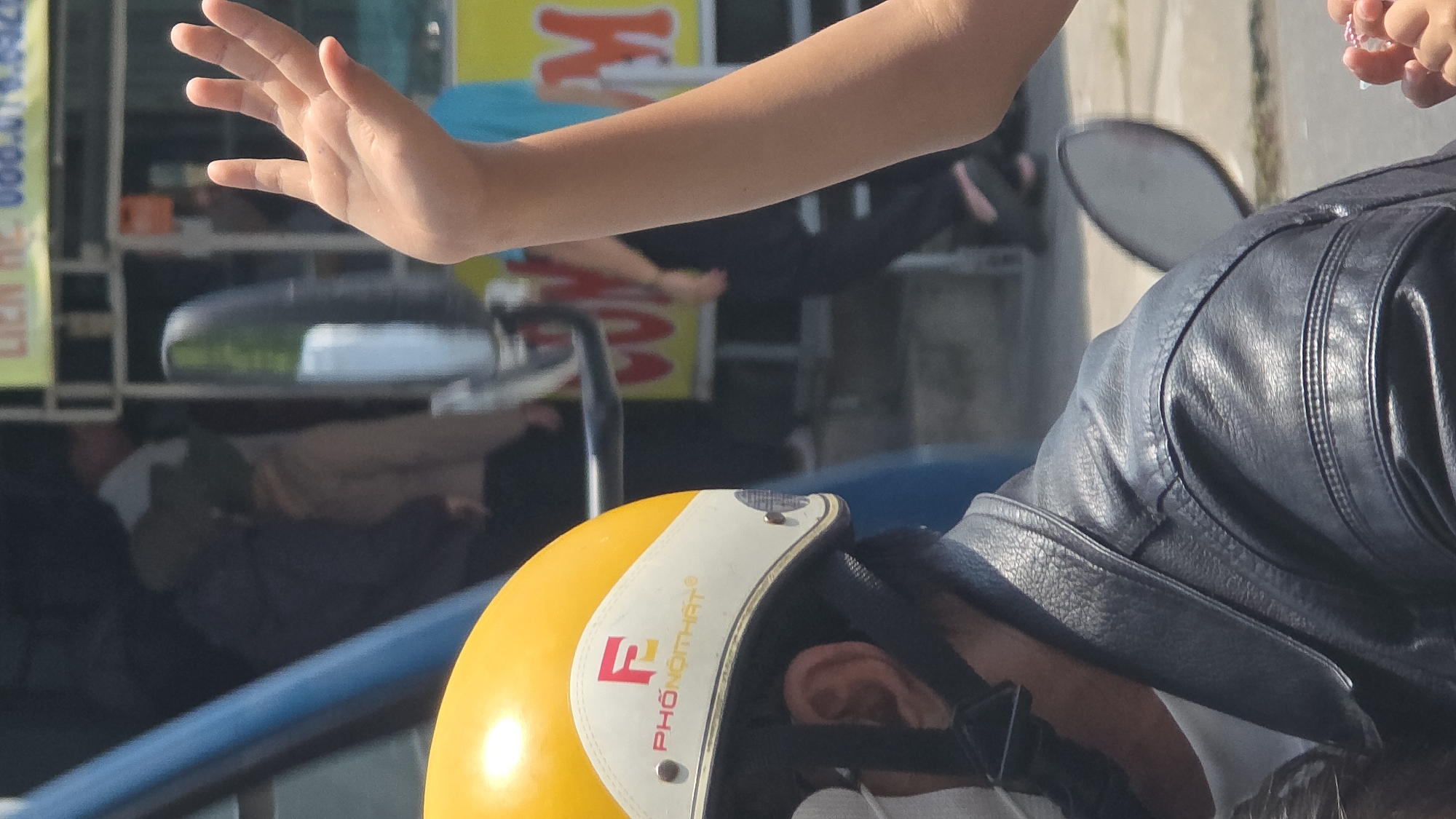 Captured from a high angle in Nha Trang, Vietnam, the image presents a dynamic street scene. A child's hand is prominently displayed in the foreground, palm facing forward. Adjacent to this, an adult wearing a black leather jacket and a yellow helmet labeled "PHO NOT HAT" is visible, suggesting they are riding a motorbike. In the background, pedestrians walk along the street near parked motorbikes and a storefront displaying signage. The environment is typical of an urban area in Vietnam, characterized by busy streets and a mix of transport and commerce.