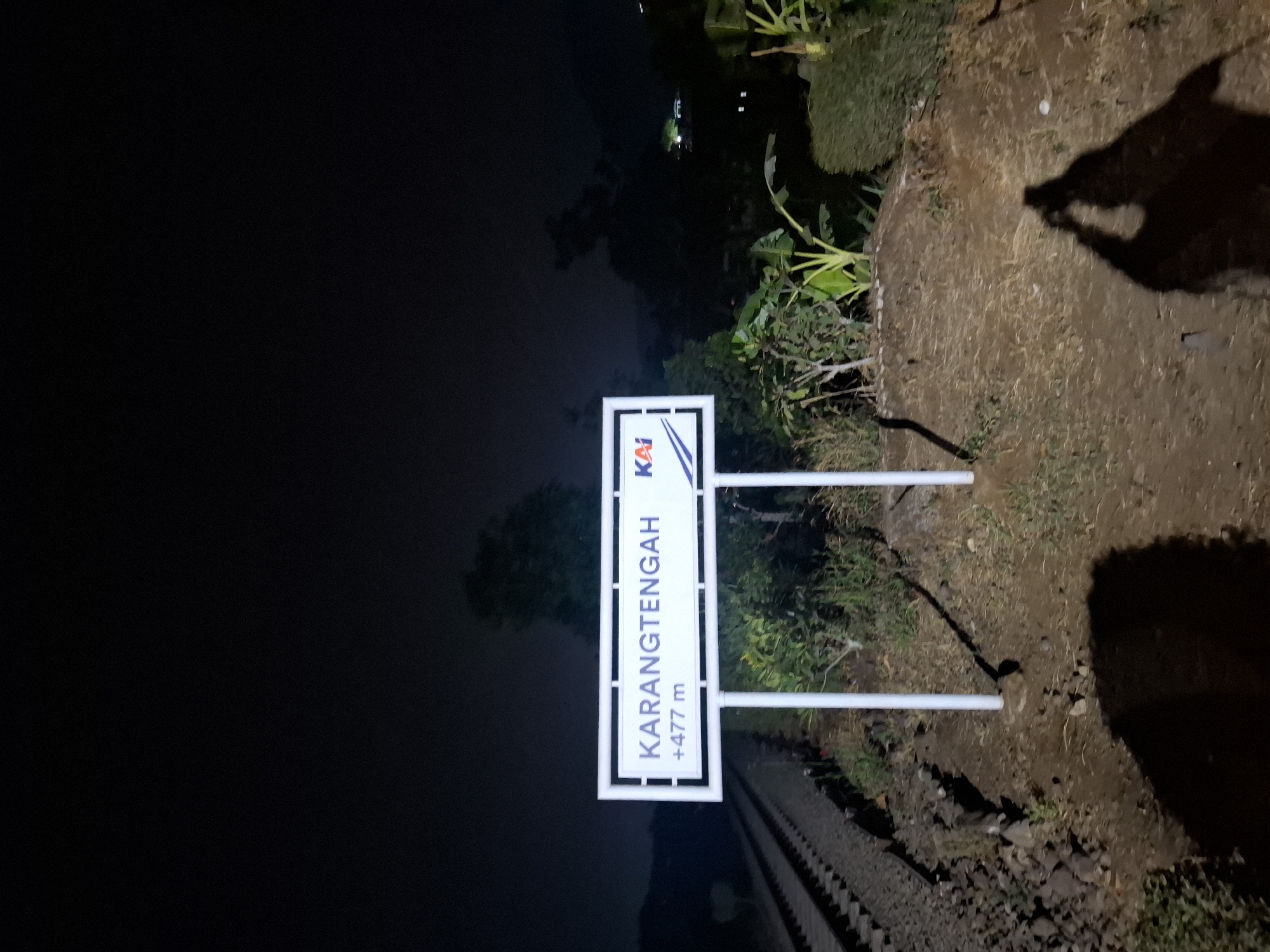 A nighttime outdoor scene captures a white sign with blue text indicating "KARANGTENGAH" and "+477 m", along with a "KAI" logo. The sign is mounted on two white posts. To the left of the sign, railroad tracks are visible, extending into the darkness. The ground surrounding the sign is covered in dirt and dry grass. In the background, a dark, hilly landscape is present, with some distant lights suggesting structures. The shadows of two people are cast on the ground in the foreground, indicating their presence at the time the photograph was taken. The overall environment appears to be rural or on the outskirts of a populated area.