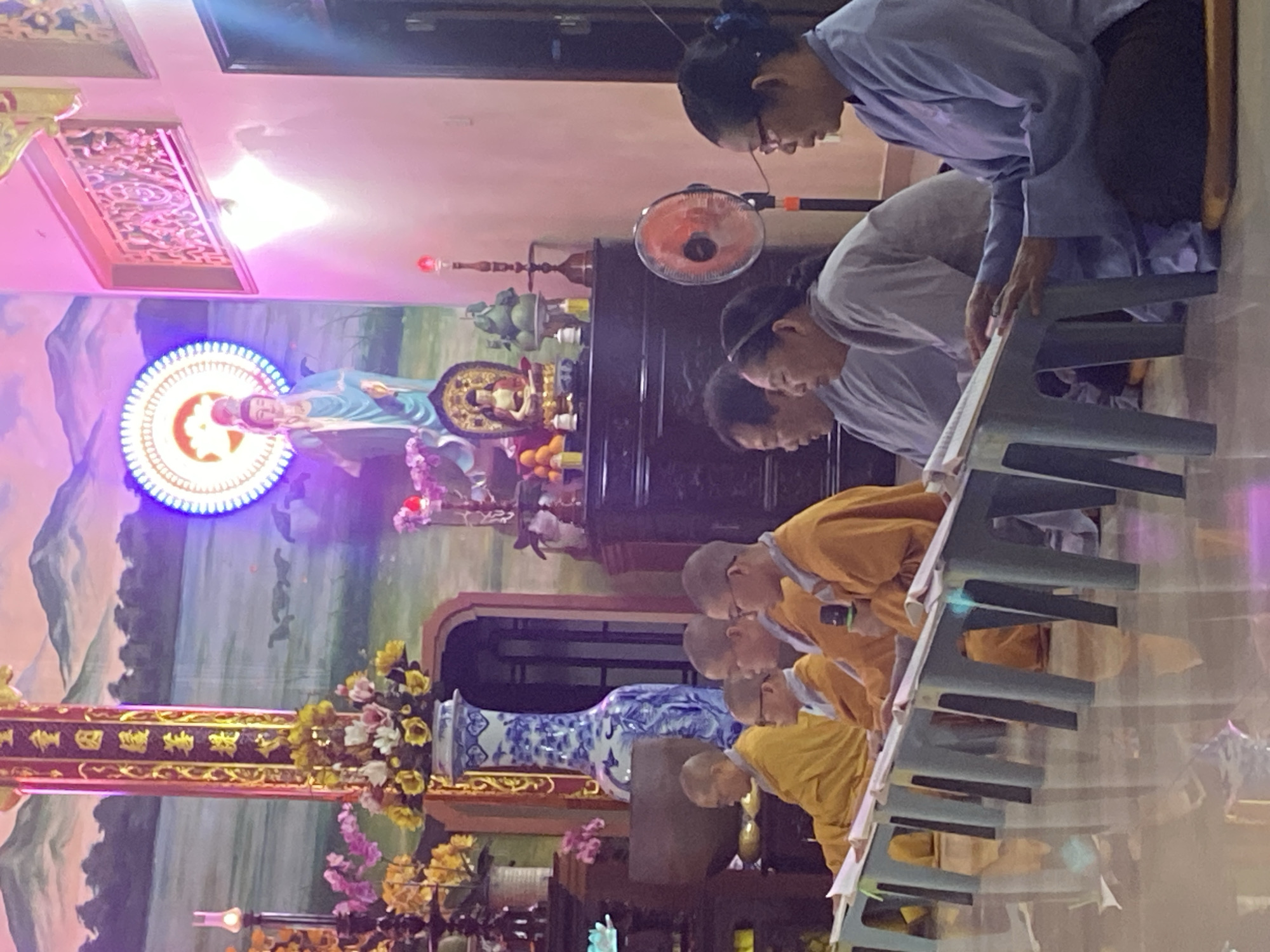 The image depicts an indoor scene within a Buddhist temple in Phu Thuy Ward, Vietnam. In the foreground, a group of individuals, likely monks or nuns based on their attire, are seated on the floor, facing forward. They are dressed in saffron-colored robes and some have shaved heads. Each person is positioned behind a light-colored plastic table, upon which they are reading or studying books. One individual in the center is holding a microphone.
The background features a large mural or tapestry depicting a mountainous landscape under a purple sky. A statue of a serene figure, possibly a deity, stands prominently against a circular, illuminated halo. Various decorative elements, including a large blue and white vase, floral arrangements, and smaller statues or offerings, are visible on shelves and furniture. A standing fan is positioned to the right of the seated individuals. The overall lighting suggests an artificial, perhaps colorful, illumination within the temple. The floor appears to be polished and reflects the light and some of the objects in the room.