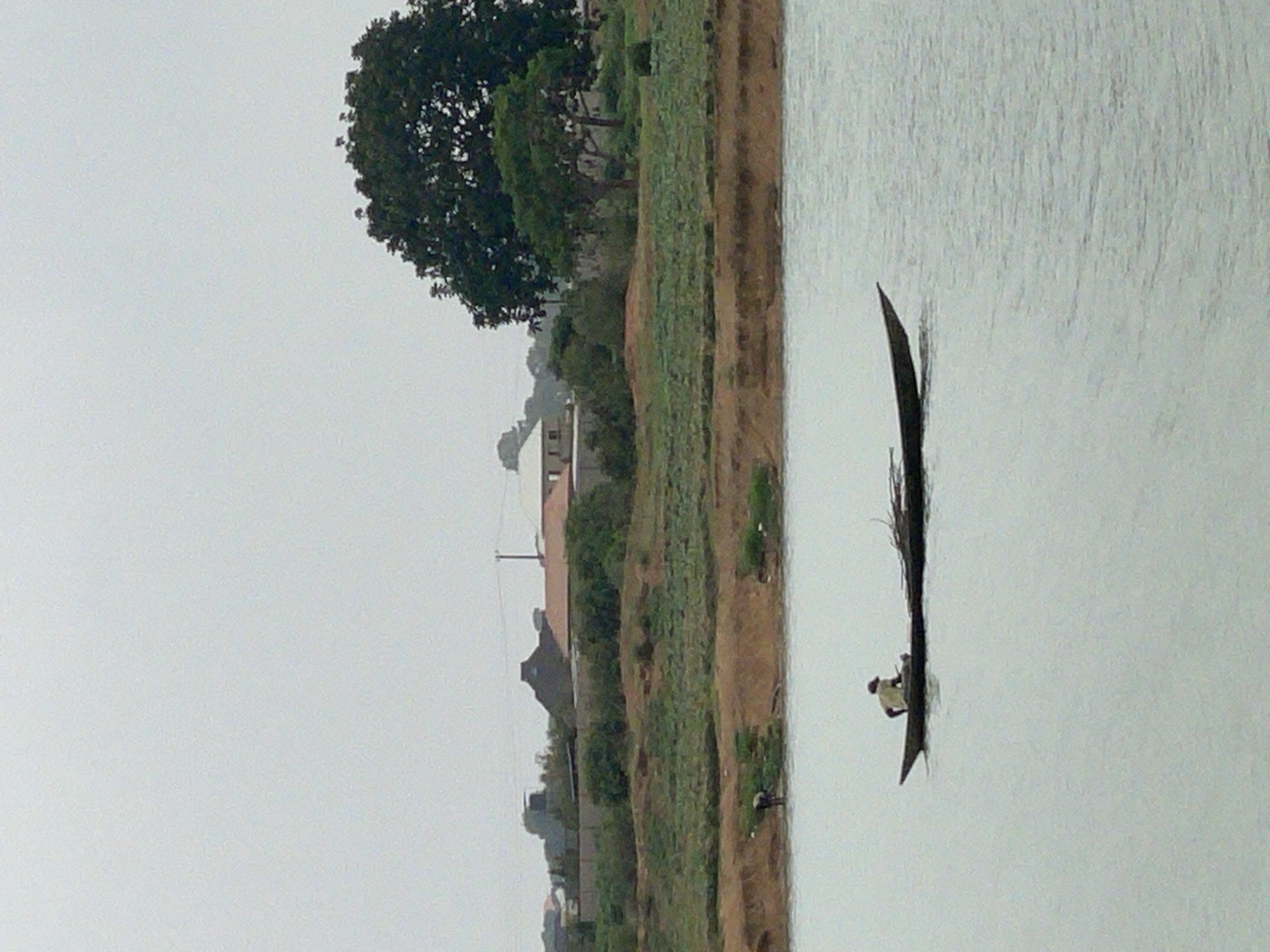 A man in a narrow boat is on the water, next to a grassy bank and some buildings, in Nabatum, Nigeria.