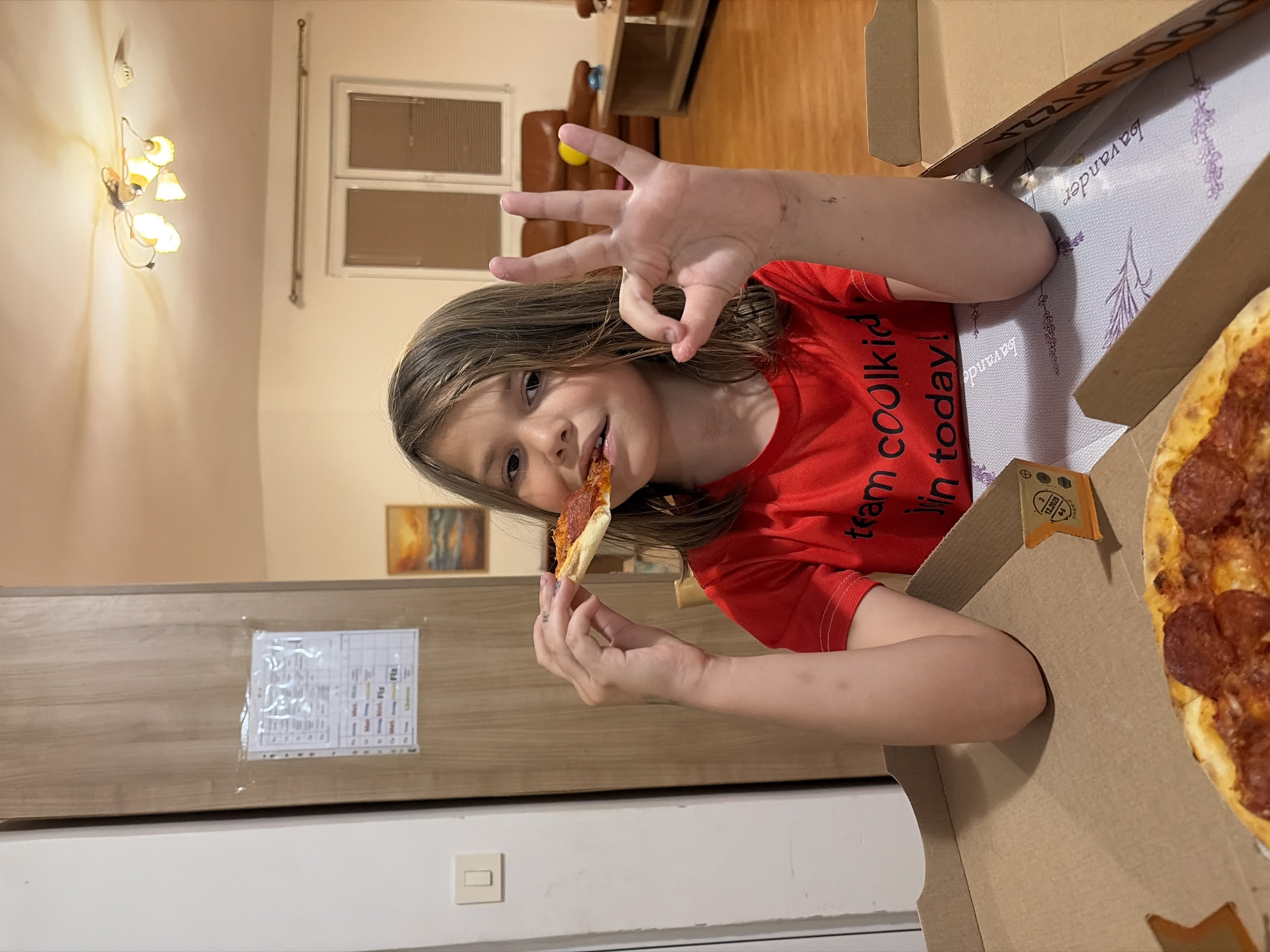 A young person with long, light brown hair is positioned adjacent to an open pizza box containing a pepperoni pizza. The child is wearing a red T-shirt with writing and eating a slice of pizza while making an "OK" gesture with their hand. The scene is indoors, with elements such as a light fixture, a window, a brown leather sofa, and a piece of artwork visible in the background. The lower portion of the image shows a section of a light brown cabinet or wall with a diagram affixed. The image was taken in a domestic environment, with no indication of its location being in Budva, Montenegro or any other specific city or place.