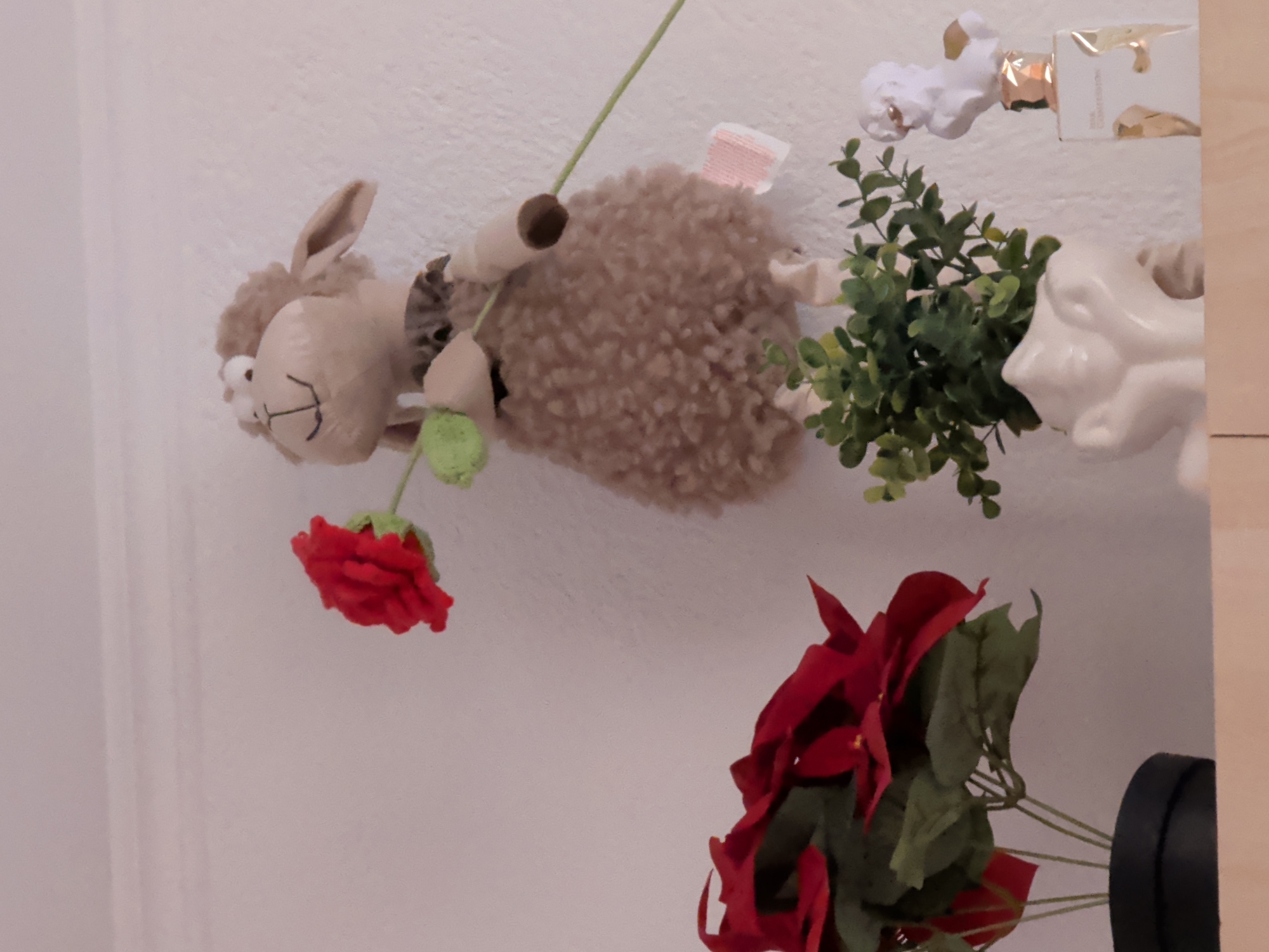The image depicts a close-up, eye-level shot of a shelf with decorative items. On the left, a black pot contains a faux poinsettia plant with vibrant red leaves and green foliage. To the right of the plant, a stuffed sheep toy with a fluffy brown body and a light brown head is holding a red flower with a green stem. The sheep is positioned on the shelf, with its body partially obscuring the stem of the flower. Further to the right, a small white pot holds a green artificial plant. Next to the green plant is a white bust figurine with gold accents, resting on a white and gold rectangular bottle that appears to be a perfume bottle. The items are arranged against a white textured wall. There is no visible indication of the city of Saedinenie, Bulgaria in the image.