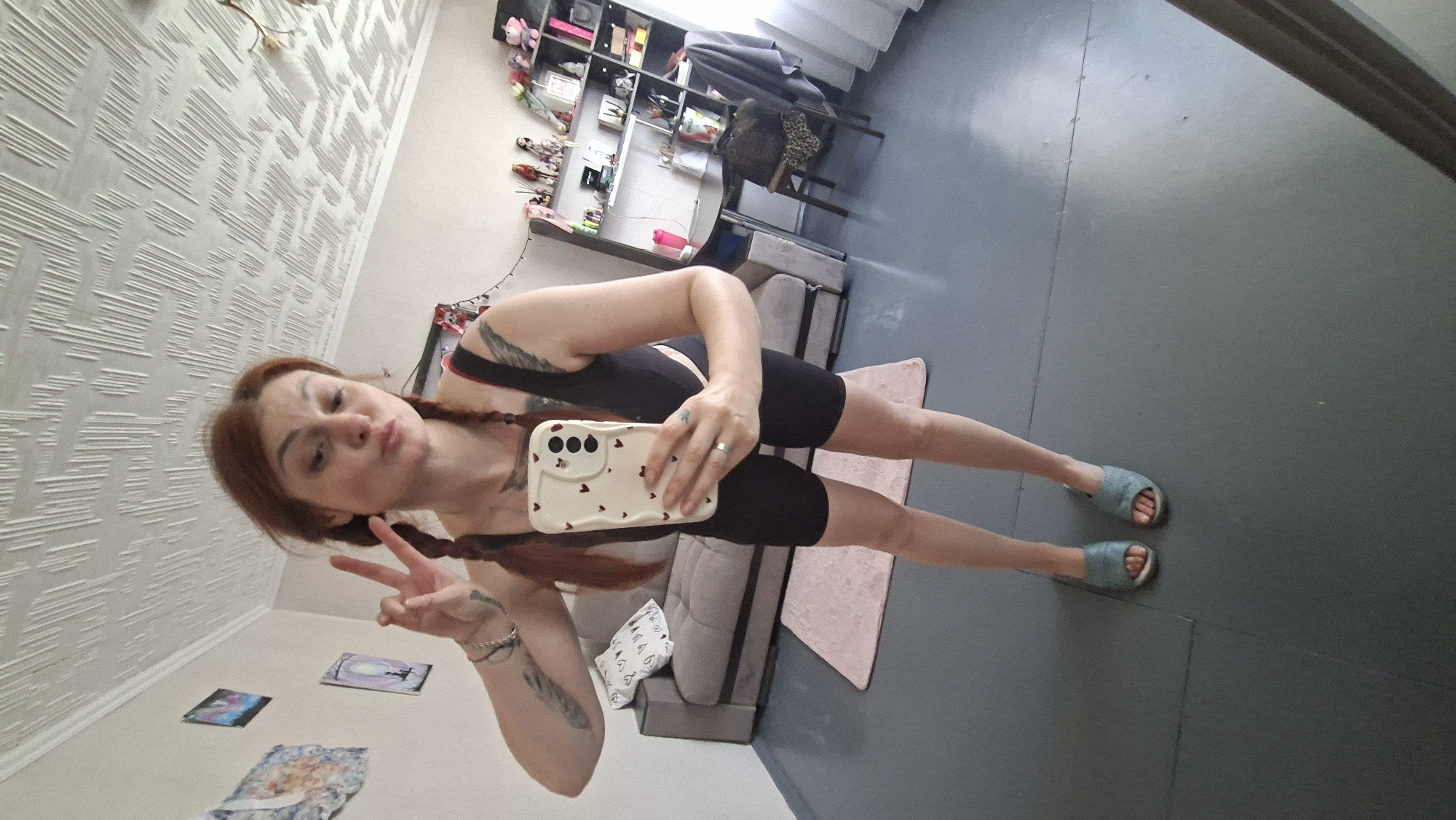 A woman with auburn hair in two braids is taking a mirror selfie in a room. She is wearing a black sleeveless top and black biker shorts. She is also wearing light blue slide sandals. She is holding a phone in her right hand and making a peace sign with her left hand. In the background, there is a grey sofa with a pink rug in front of it. To the right, there is a bookshelf filled with various items, including dolls and decorative objects. The room has a textured ceiling and light-colored walls. The floor is grey. The image appears to have been taken in a residential setting.