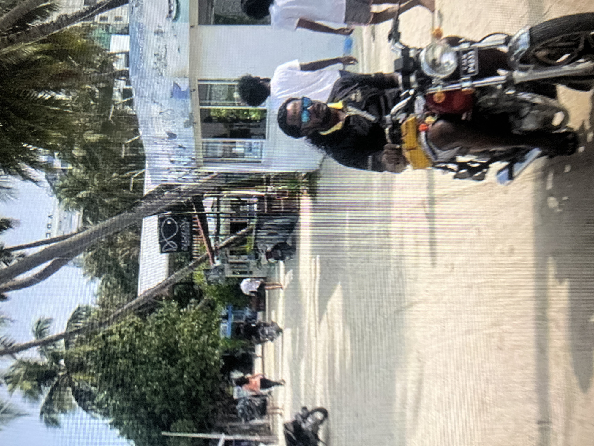 A man with long dark hair and blue sunglasses rides a motorcycle on a sandy path lined with palm trees and buildings. He is wearing a black shirt with yellow accents and white shorts. In the background, several people are walking and riding motorcycles. There are shops and buildings along the path, and the sky is bright and hazy. The scene appears to be a tropical or subtropical location. The city of Qarağandı city administration, Kazakhstan is not depicted in this image.