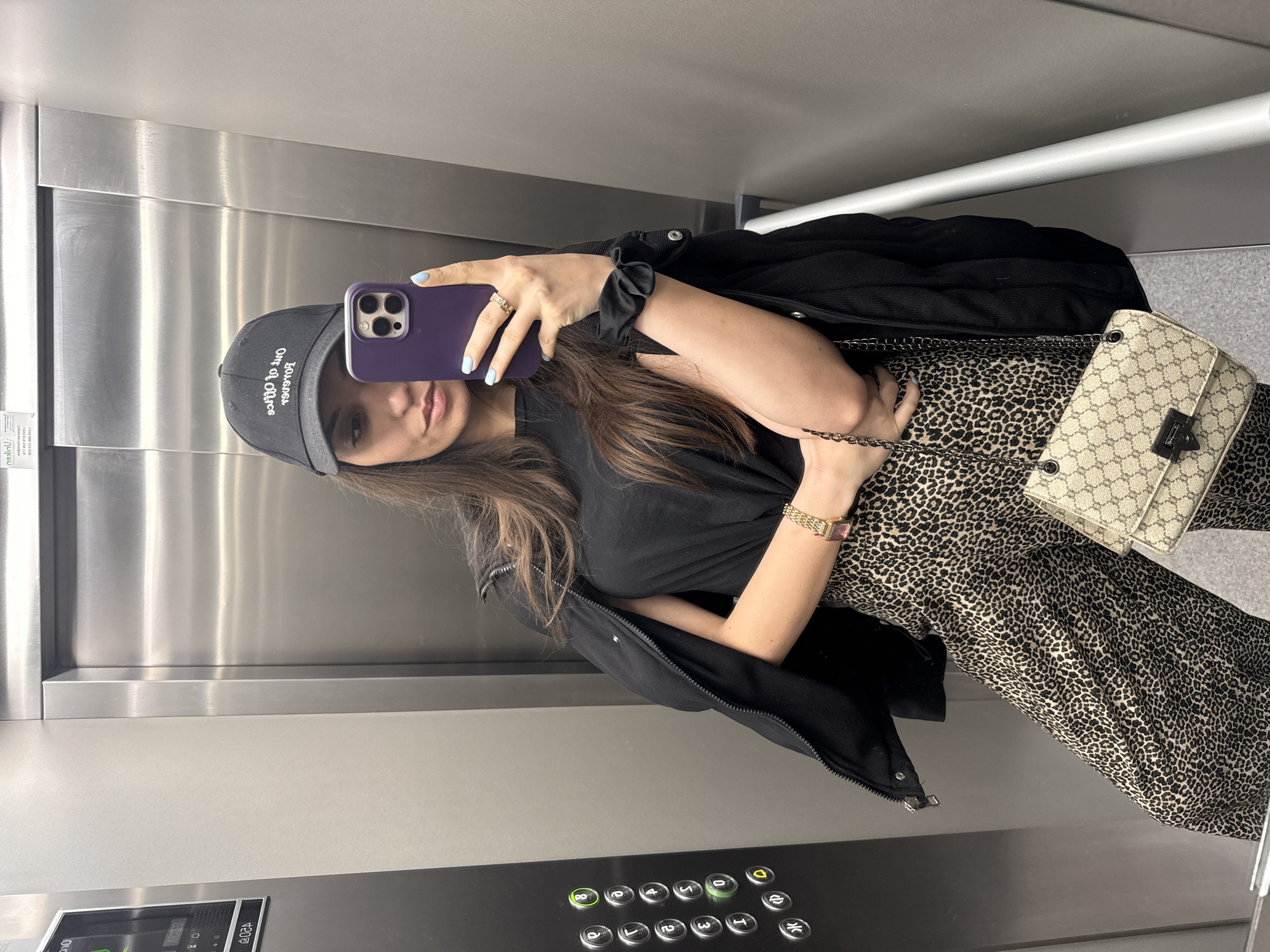A woman is taking a selfie in what appears to be an elevator. She is wearing a black cap with white writing, a black t-shirt, a black jacket, and a leopard-print dress or pants. She also has a gold-colored watch on her left wrist and a black scrunchie around her right wrist. She's holding a purple phone in her right hand, and a beige purse with a chain strap hangs on her right side. The elevator has brushed metal walls and a panel of elevator buttons. No specific details indicate it is in Plovdiv, Bulgaria.