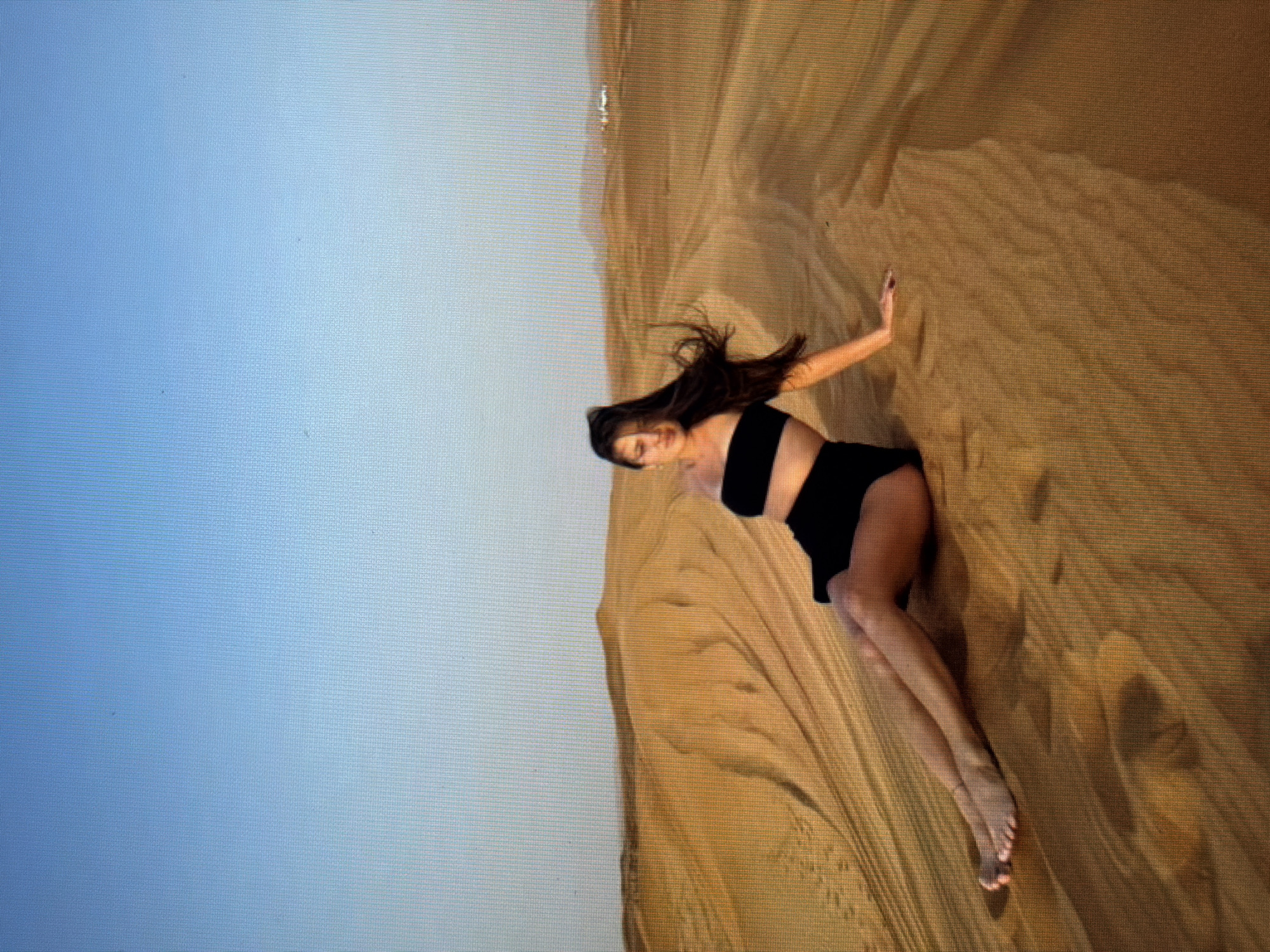 A young woman with long, dark hair is sitting on a sand dune in the desert. She is wearing a black bandeau top and black shorts. Her legs are extended in front of her, and she is leaning back on her left hand. The sand dunes stretch out behind her, with a clear blue sky above. There are no other people or objects visible in the image, except for some distant buildings on the horizon. The image appears to be taken during the day, possibly at sunset, given the warm lighting on the sand. The location of Sirius, Russia is not depicted in this image.
