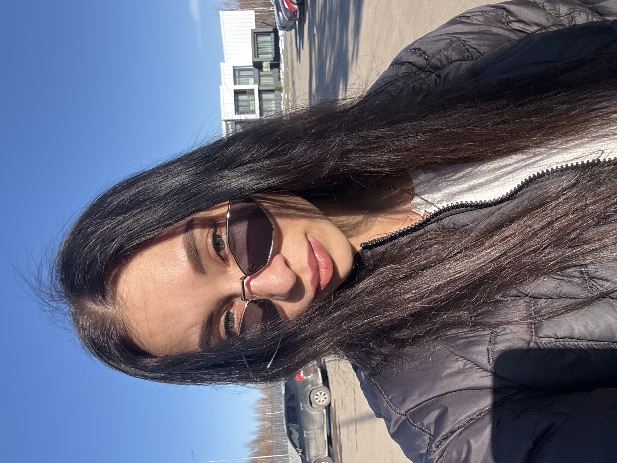A young woman is photographed outdoors on a sunny day. She wears sunglasses, a white shirt with a partially zipped jacket, and has long dark hair. The background includes modern residential buildings and parked cars, suggesting an urban environment. While the location is identified as the city of Votkinsk, Russia, there are no visible landmarks or context to definitively confirm the city.