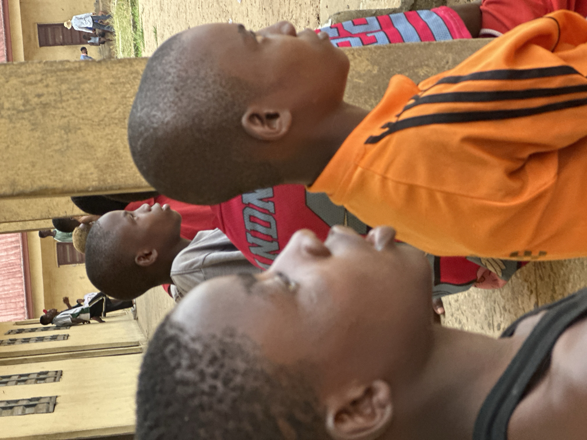 An outdoor scene in Ikot Ekpene, Nigeria, depicts multiple individuals, primarily youth, near a light yellow-tan building with a reddish-brown corrugated metal roof. The foreground features three dark-skinned males. The closest, on the left, is partially visible from the left side of his head and upper shoulder; he wears a dark grey or black tank top and looks towards the right. Immediately behind him and to the right, a second male youth is in profile, facing right, wearing an orange long-sleeved shirt with three black vertical stripes on the right sleeve. His gaze is directed forward and slightly downward. Behind him, a third male youth is seen from the chest up, wearing a red shirt with white lettering.
In the midground, a light brown/tan column or wall section partially obscures the background. To the left of this column, a light yellow-tan building with multiple rectangular shuttered windows has a covered walkway. Under this walkway, a male figure wearing a white t-shirt, dark shorts, and a red cap leans against the building wall, looking left. Further back, another figure is partially visible under the walkway.
The ground is composed of dirt or sand with sparse patches of green grass. In the background, another similar light yellow-tan building with a reddish-brown corrugated metal roof and a dark door is visible. On the dirt ground to the far right, a male figure wearing a light-colored long-sleeved shirt and a straw hat stands, looking left. Near the second building, another figure sits on a low object wearing a white tank top, while another is partially visible bending over. The overall scene suggests a community or schoolyard environment under daylight conditions.