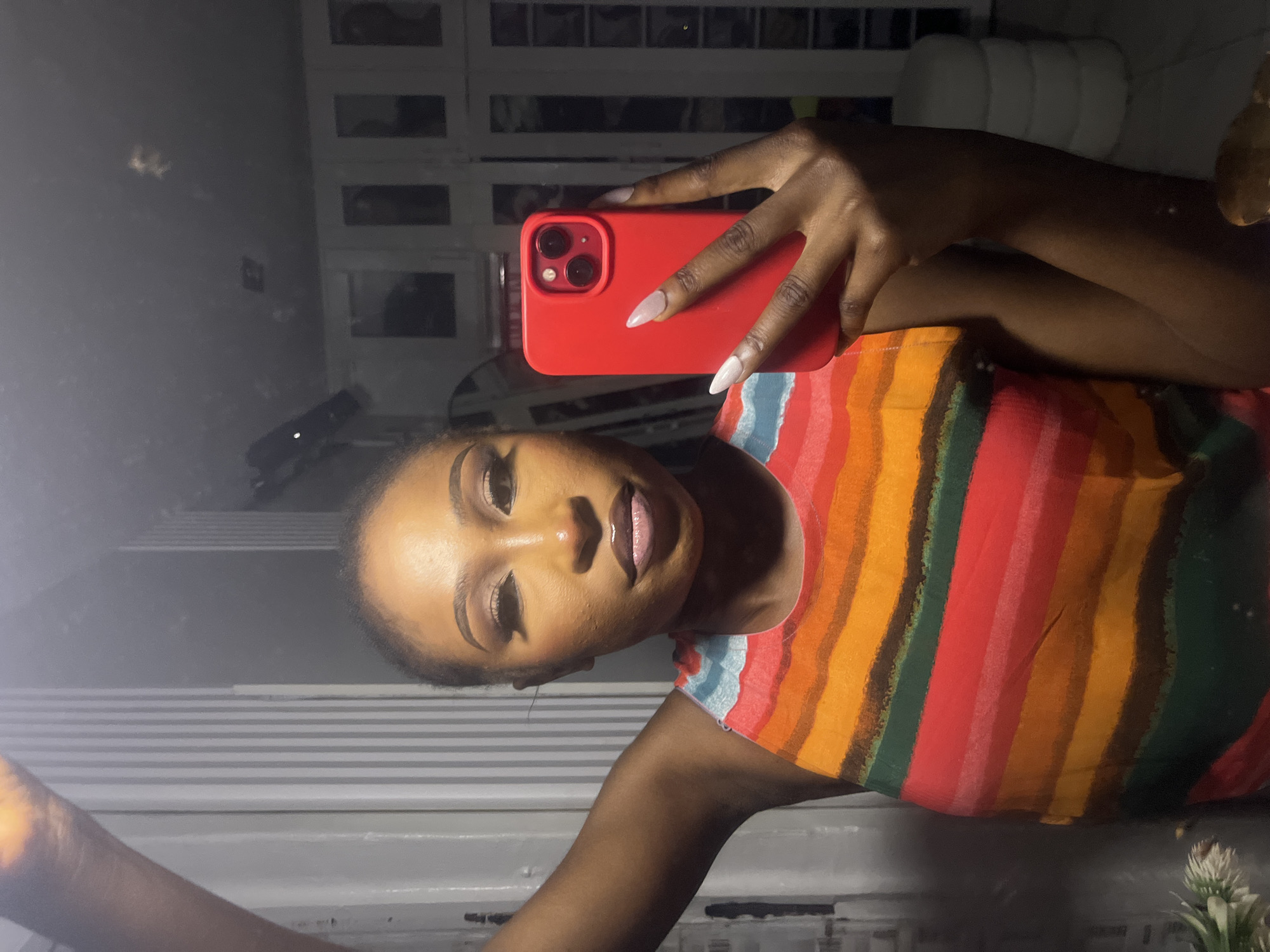 A young African woman takes a selfie in a brightly lit indoor setting, possibly a bedroom or dressing room. She is wearing a sleeveless top with bold horizontal stripes in red, orange, yellow, green, and brown. Her right arm is raised, holding a red smartphone, which is angled to capture her reflection. Her makeup is meticulously applied, with defined eyebrows, winged eyeliner, and a glossy lip color. Her nails are long and painted a light color. In the background, vertical blinds are visible, along with a mirrored closet or wardrobe. The overall impression is one of stylish self-presentation within a modern domestic space, likely within the city of Abuja, Nigeria.