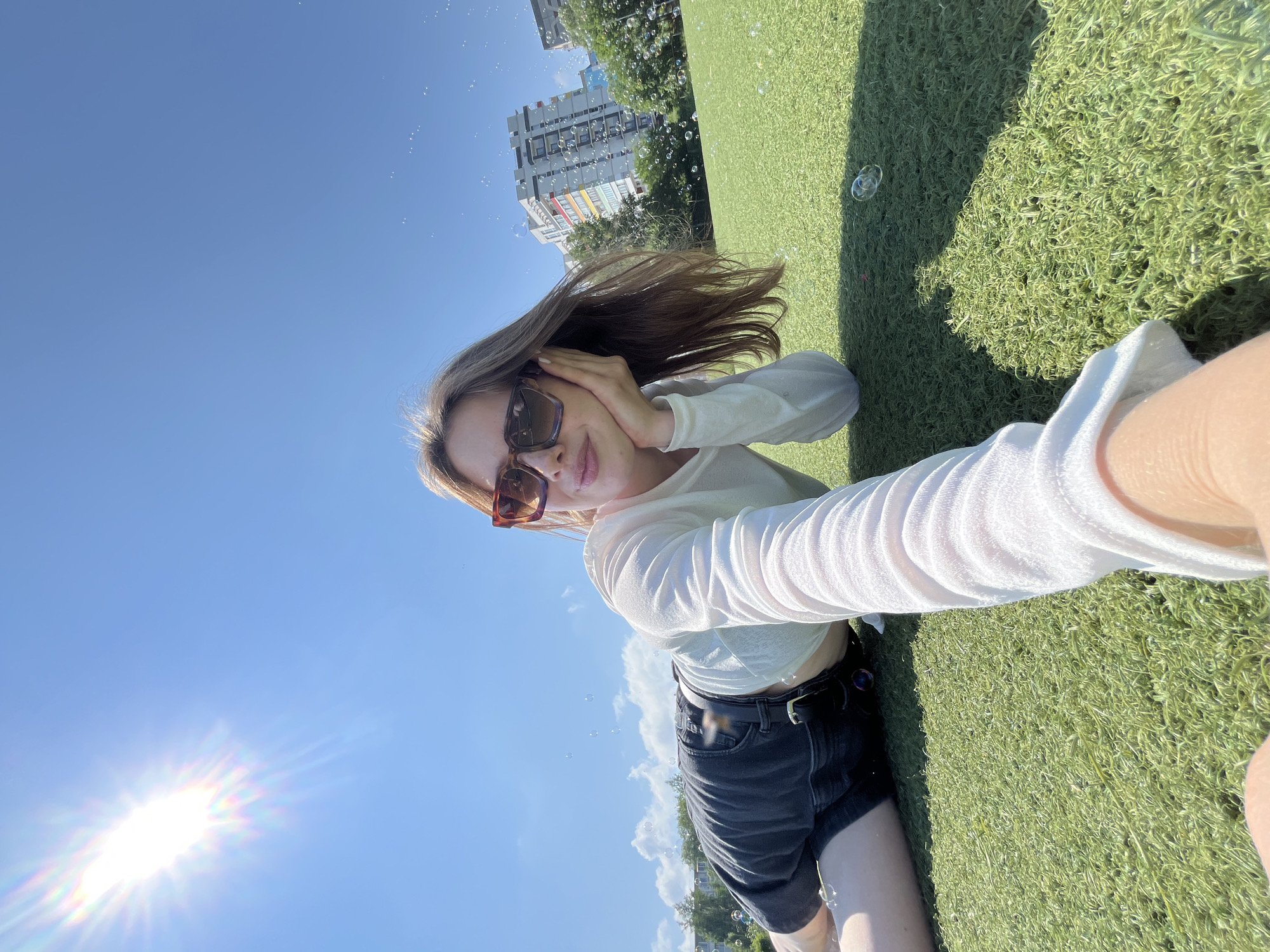 A young woman with brown hair, wearing sunglasses, a white long-sleeved shirt, and black shorts, lies on a grassy field. She is holding her head with one hand and taking a selfie with the other. In the background, buildings and trees are visible. Bubbles are floating in the air. The sun is shining brightly in a clear blue sky. The location is likely a park or recreational area in Chaikovsky, Russia, given the urban buildings in the distance and the well-maintained grassy field.