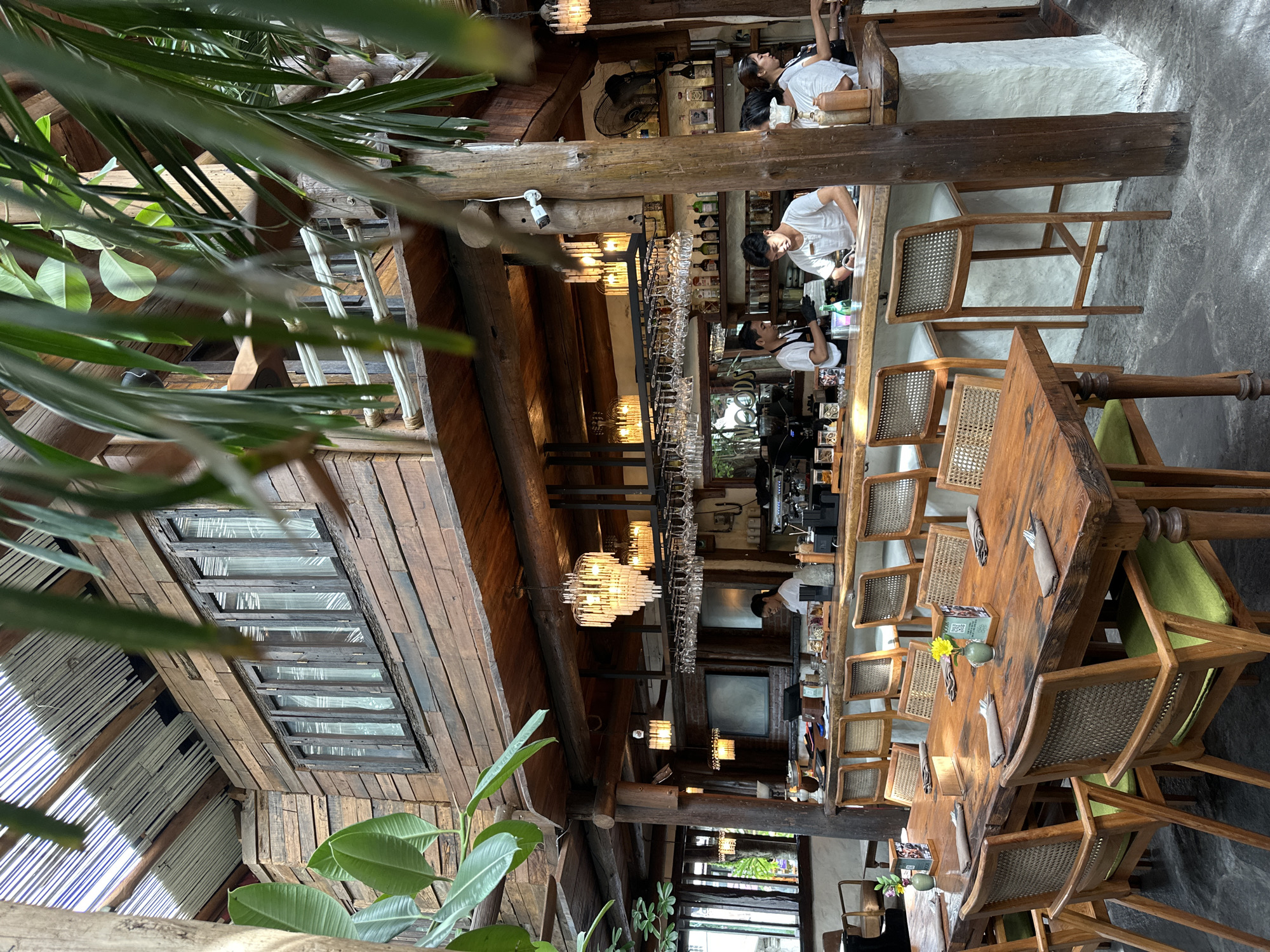Captured in Mengwi, Indonesia, the image depicts the interior of a restaurant. The restaurant has a distinctive architectural style that is a mix of rustic and modern elements. The roof, crafted from wooden planks, includes a section with windows to allow natural light to filter in. A variety of lighting fixtures, including chandeliers and smaller decorative lights, enhance the ambiance.
A long wooden table with chairs is a prominent feature, suggesting a communal dining experience. Place settings on the table indicate it is prepared for customers. Behind the table is a bar area where staff members are present. One staff member is actively engaged behind the bar, and customers are seated at the bar counter. Shelves behind the bar are stocked with bottles, presumably alcoholic beverages. The overall setting is detailed and indicative of a well-maintained and inviting space. Green foliage both inside and outside contributes to a tropical atmosphere.