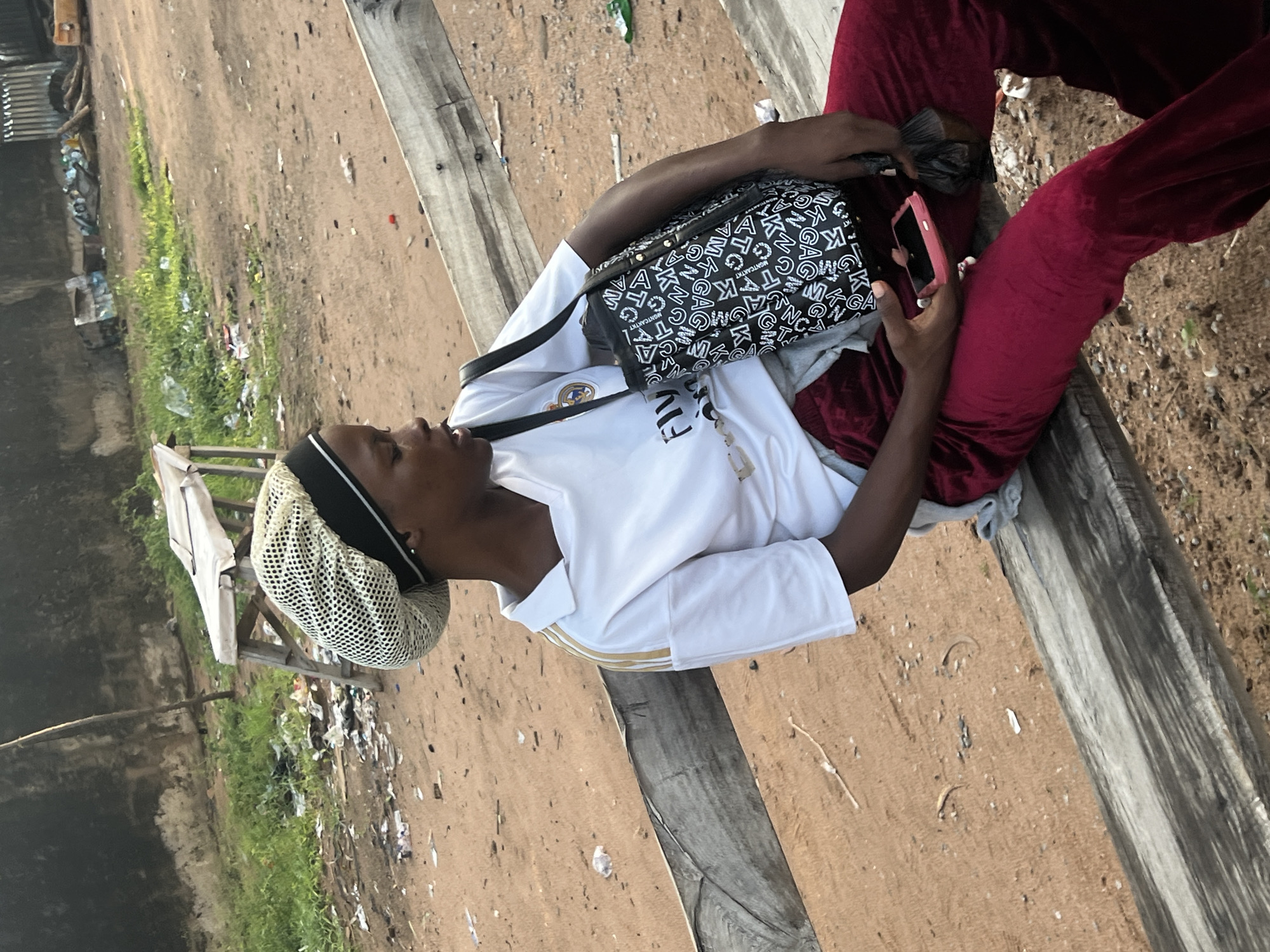 A young female, with dark skin, sits on a worn wooden bench, gazing to her right. She wears a white Real Madrid short-sleeved soccer jersey with gold trim and "Fly Emirates" sponsorship, over which a grey garment is visible at the waist. Her head is covered by a light-colored mesh cap with a black band. She is dressed in red, possibly velvet, wide-leg trousers. A black shoulder bag with a repeating white letter pattern hangs from her left shoulder. Her right hand holds a pink smartphone, while her left hand grips a small black plastic bag. The ground is reddish-brown dirt, characteristic of laterite soil, heavily littered with various refuse including plastic and paper. In the background, there are patches of green vegetation, a rustic wooden table with a white cloth, and a dark, weathered wall. Further back on the right, additional debris and a partial yellow object are visible. The scene depicts an outdoor, undeveloped area in Gbaza, Nigeria.