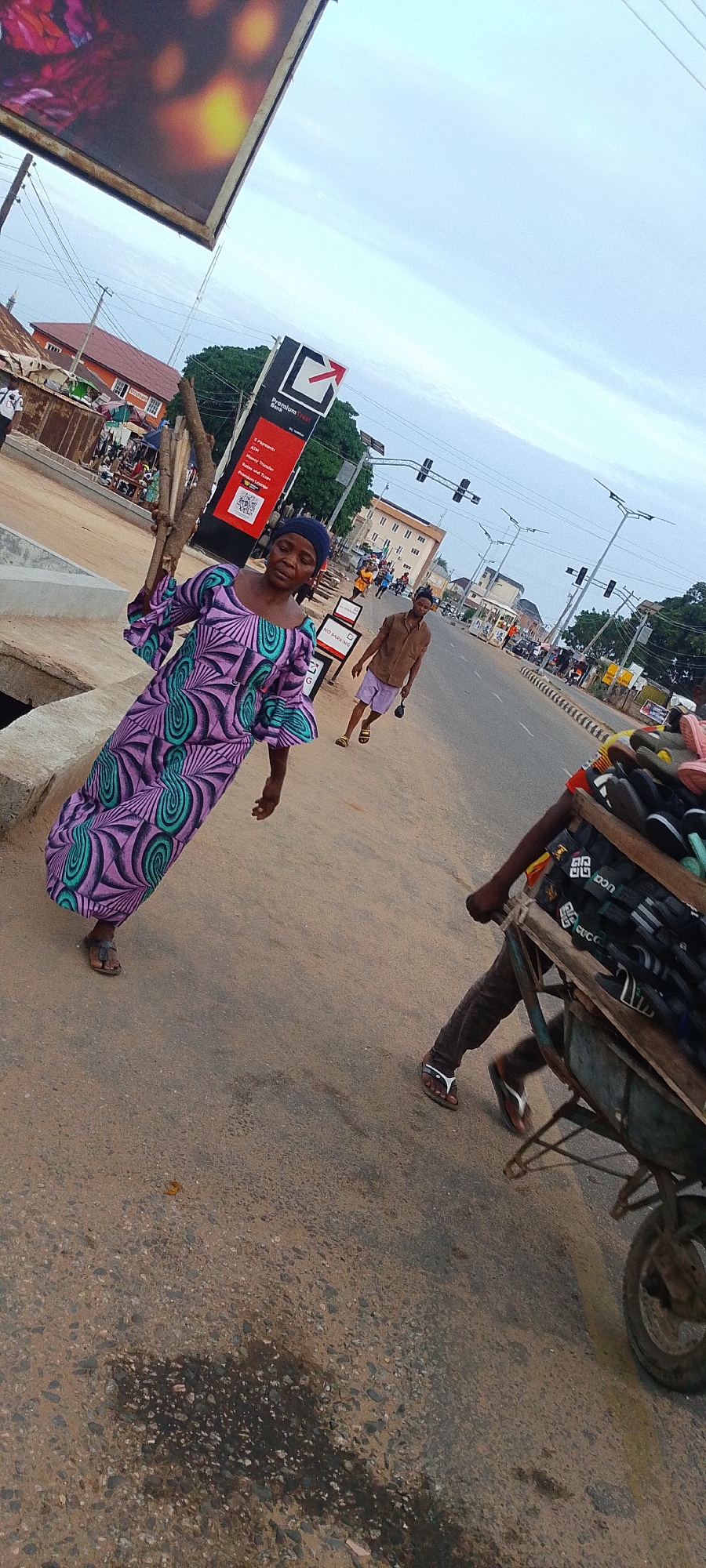 The image depicts a street scene in Lokoja, Nigeria. A woman wearing a purple and green patterned dress and a blue head covering stands on the side of the road, holding sticks. A man in a brown shirt and purple shorts walks further down the road, holding a long object. Another man pushes a wheelbarrow filled with shoes past the camera. In the background, there are buildings, traffic lights, a Premium Trust Bank sign, and other people. The street is paved and appears to be moderately busy.