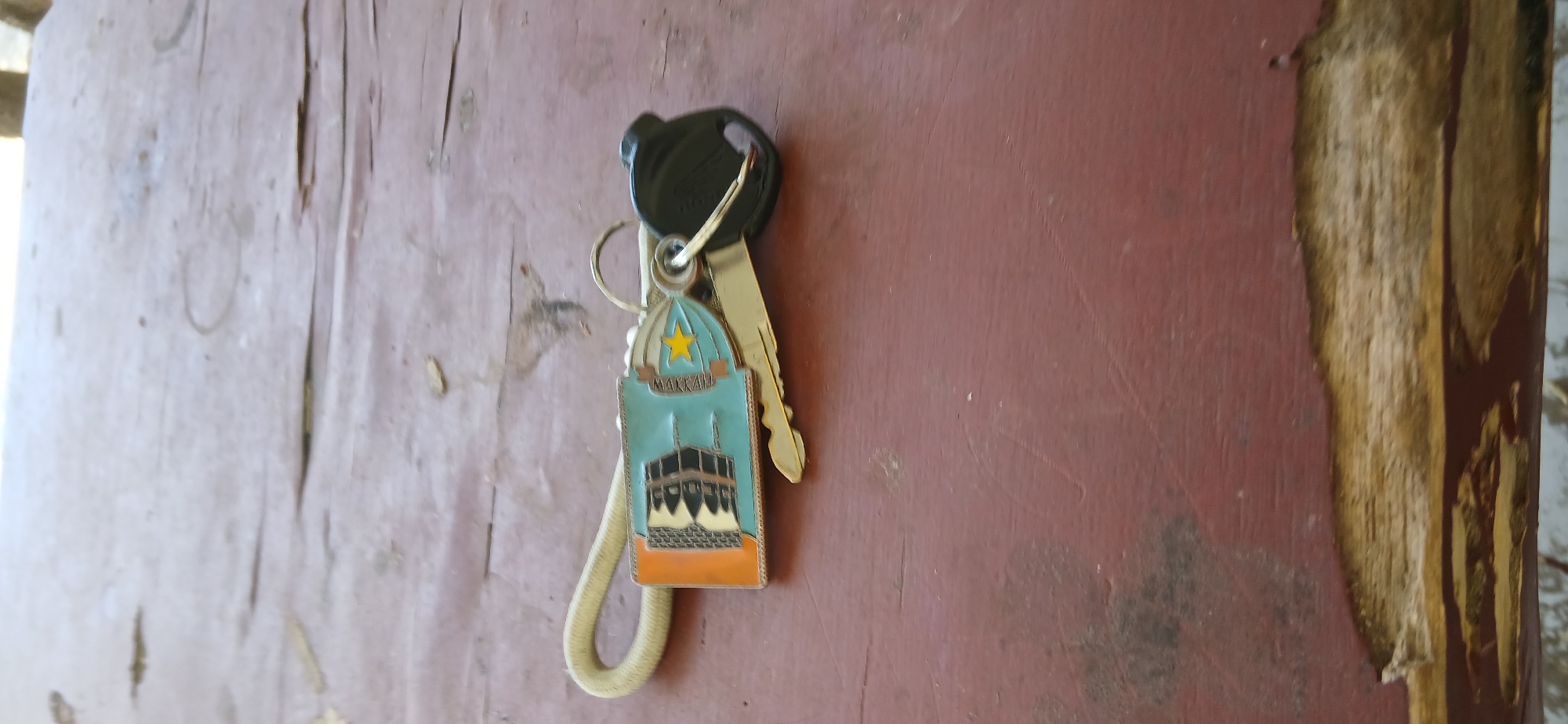 A keychain with a charm depicting a blue building with an orange border and a yellow star is attached to two keys on a worn, red wooden surface. The charm has the word "MAKKAH" written on it. One key is brass-colored and partially visible, while the other has a black plastic head. The wooden surface exhibits signs of wear, with peeling paint and exposed wood grain. The image is taken from a slightly elevated perspective, with the background out of focus and appearing to be outdoors. There is no indication of the specific location within Pejaten, Indonesia.