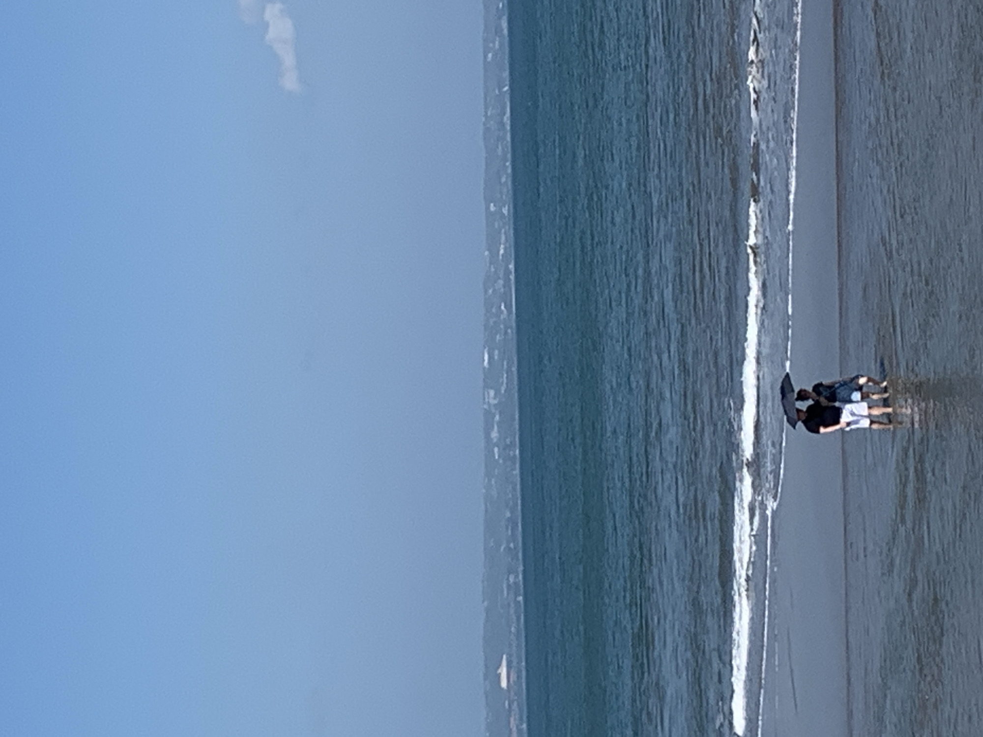 In the foreground, a couple stands in shallow water on a beach, holding a black umbrella above them. The man, on the left, wears a black t-shirt and white shorts. The woman, on the right, is wearing a blue dress. They are walking towards the right of the frame. Gentle waves lap at the shore, and the sand appears wet. The middle ground is occupied by the deep blue ocean, stretching towards the horizon. In the background, a hazy landmass is visible, with buildings and a light-colored structure, possibly a boat or a building on a promontory, to the far left. The sky above is a clear, light blue with a faint cloud on the far right. The scene suggests a beach in North Kuta, Indonesia, likely during a mild weather day.
