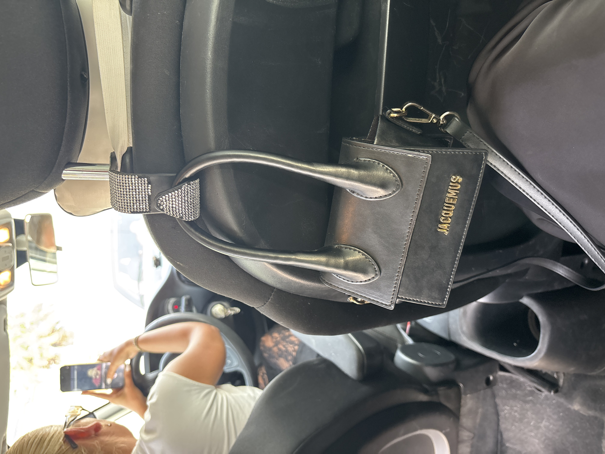 The image shows the interior of a car, focusing on a black Jacquemus handbag hanging from a car headrest hook. The hook is adorned with rhinestones. In the background, a woman with blonde hair pulled back is visible, wearing sunglasses and a white t-shirt. She is holding a smartphone and appears to be taking a photo or video while driving. The car's interior is dimly lit, with sunlight filtering in from the front. The scene is likely in Orihuela, Spain, given the context of the city provided in the prompt, although there are no explicit visual cues within the image to confirm this specific location.