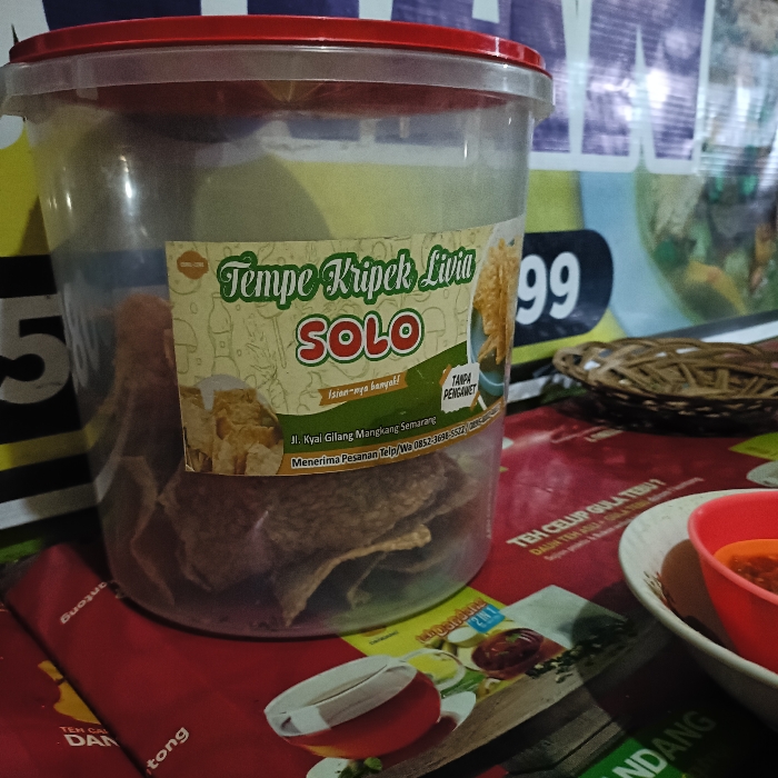 A clear plastic container with a red lid, containing a large amount of dried, flat, rectangular, brown, crispy snacks called "Tempe Kripik Livia SOLO" is situated on a red and yellow patterned table. There are other objects in the image, including a brown wicker basket, a small red bowl containing a red liquid, and a white bowl in the lower right corner of the image. The container is labeled with text in Indonesian, likely describing the product and the location and contact information for the manufacturer. The location is in Tugu, Indonesia, which is identified by the coordinates -6.975 latitude and 110.332 longitude. The table is in a restaurant or food stall environment. The image is likely taken from a customer's perspective.