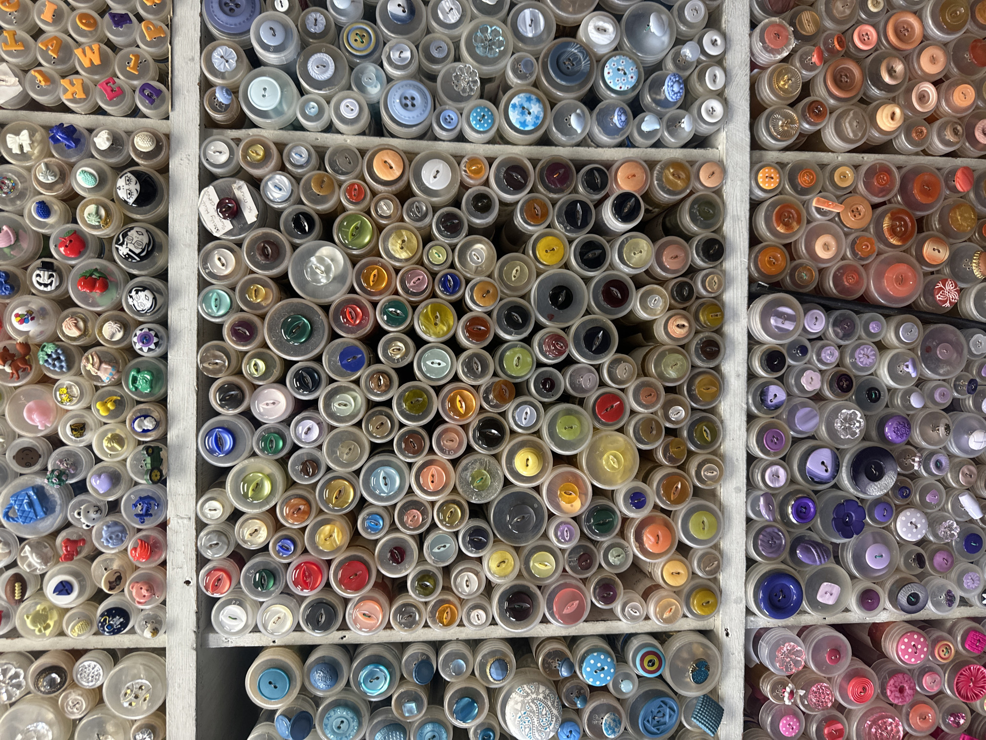 The image shows a close-up view of a collection of buttons neatly arranged in small, clear containers, organized within a set of cubbies or shelves. The buttons vary widely in color, size, and design, ranging from simple, plain buttons to more decorative ones with patterns, shapes, and embellishments. The containers are predominantly circular. The grid-like structure of the shelves and the systematic arrangement of the containers suggest an organized collection or display.
The OCR output includes text fragments such as "K", "M", "Well", "lly Hall" and "Sevenoaks" which, along with other glyphs, seem to be extracted from labels or writing possibly associated with the buttons or their containers.
Based on this image being located in Sevenoaks, United Kingdom, the collection is likely located in a crafts store or a personal collection in a home within this city.