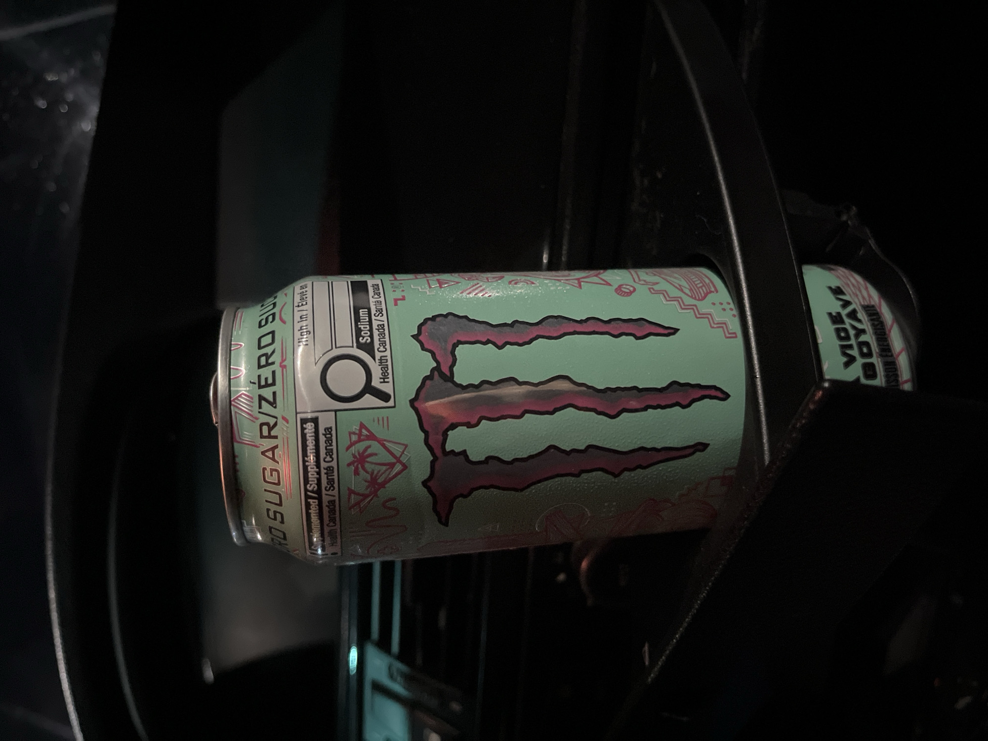 A close-up shot depicts a can of Monster Zero Sugar energy drink, specifically the "Vice Goyave" flavor. The can is held within a dark, indeterminate structure, possibly a vehicle's cup holder or compartment. The can's label shows nutrition information in both English and French, indicating compliance with Health Canada regulations, and highlights the sodium content. The background is dark and indistinct, suggesting a low-light environment. There are no people visible. There is no information to suggest this image was taken in Shumen, Bulgaria.