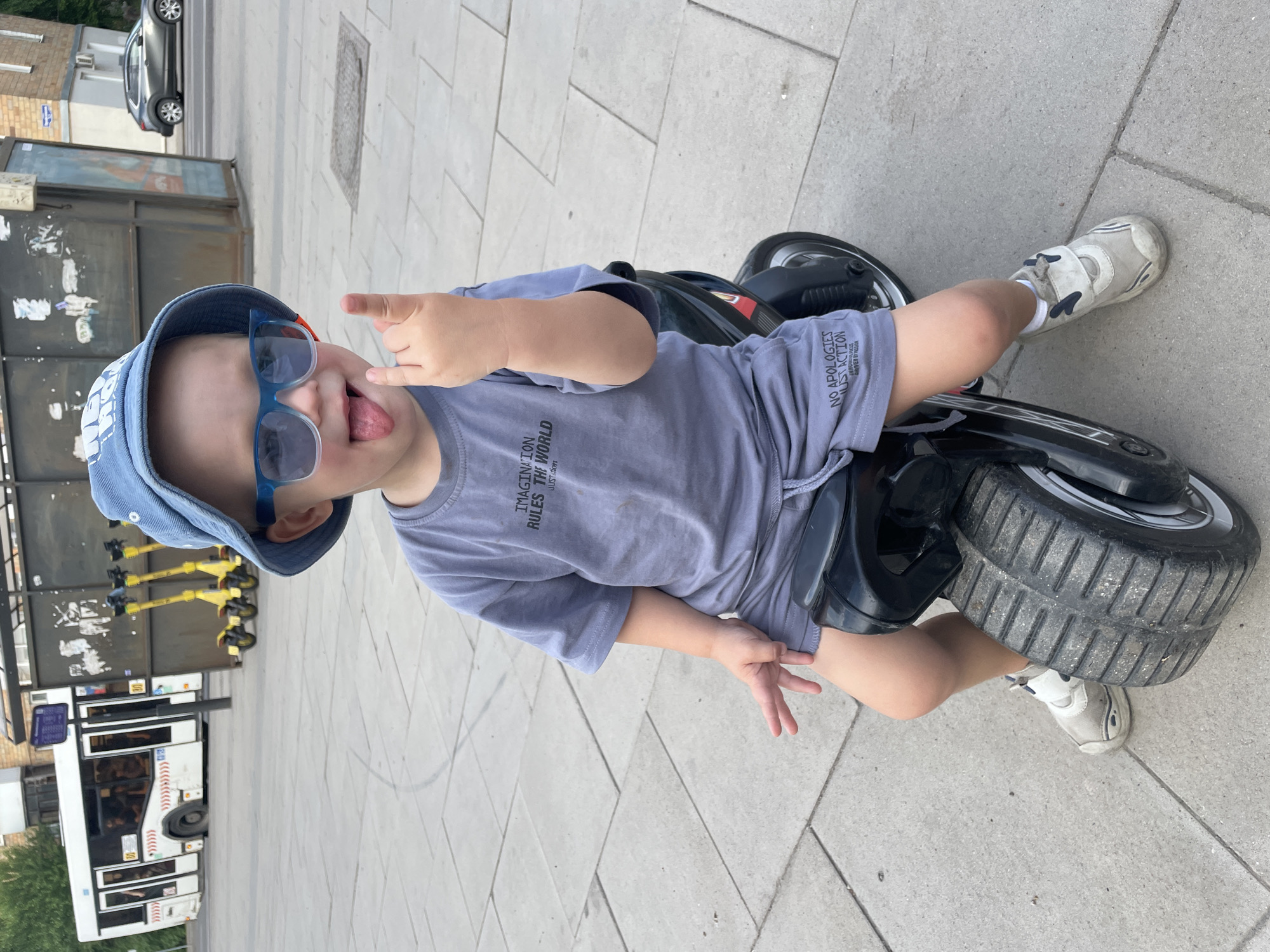 A young child wearing a blue bucket hat and blue sunglasses is sitting on a black toy motorcycle. The child has their tongue sticking out and is making a rock-and-roll hand gesture. They are wearing a grey t-shirt with the text "IMAGINATION RULES THE WORLD" and grey shorts with the text "NO APOLOGIES JUST ACTION". The child is in an outdoor urban environment with a paved sidewalk made of light grey square tiles. In the background, there is a white bus, several yellow electric scooters, and a dark grey metal structure. A dark grey car is visible on a street in the distance. The scene appears to be in an urban setting, possibly a city square or pedestrian area.