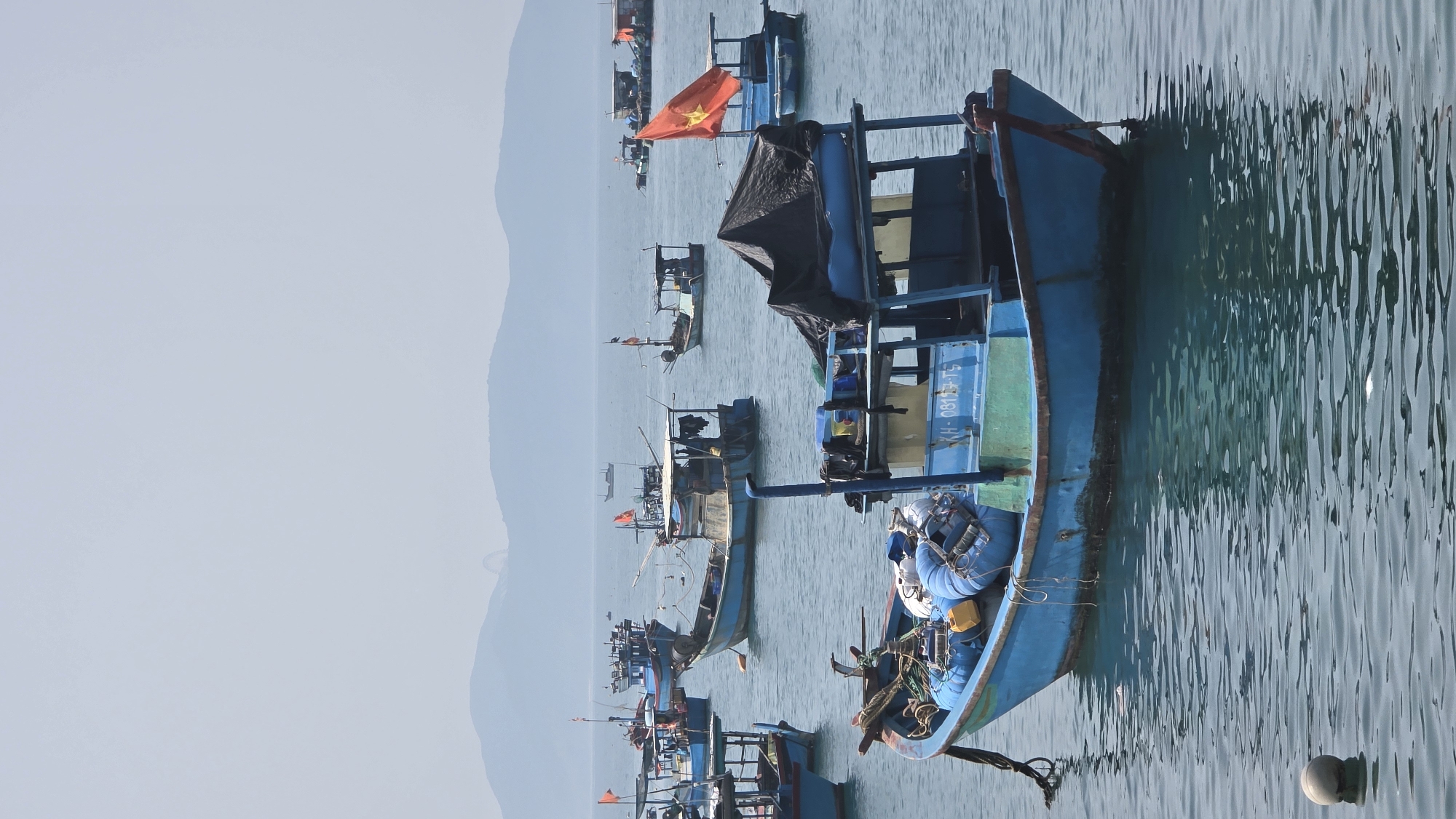 The image depicts a harbor scene in Bắc Nha Trang, Vietnam, featuring numerous blue wooden fishing boats floating on calm water. Several boats are adorned with Vietnamese flags. Mountains are visible in the background under a hazy sky. The close-up boat has gear such as ropes, tires, and blue plastic barrels, with a license plate number on its side. There is no evidence of people in the close-up view, but other boats appear to have people aboard. The water reflects the blue tones of the boats and sky.