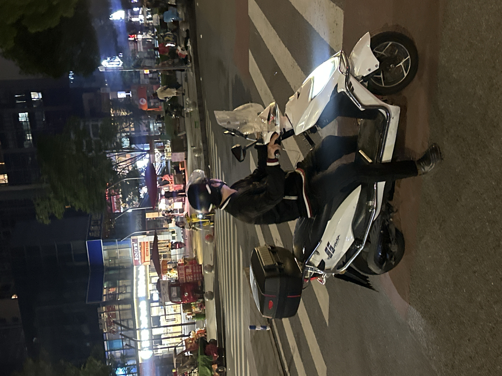 A person wearing a black helmet and black clothing is riding a white scooter at night. The scooter has a black storage box on the back and a clear windshield. The scooter is stopped at a crosswalk. In the background, there are buildings with illuminated windows and storefronts, trees, and other vehicles. The location is the city of Chongqing High-tech Zone, China.