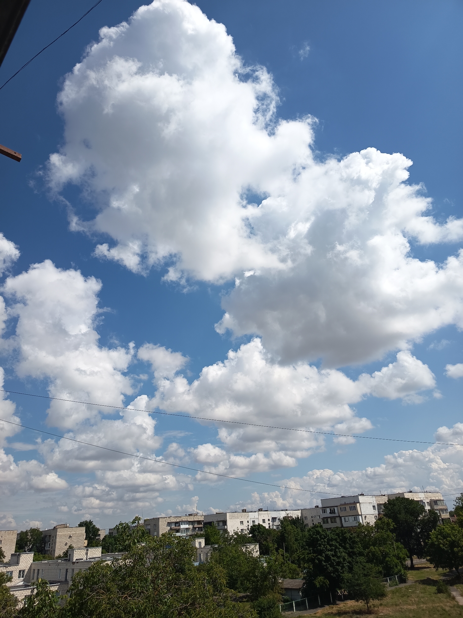 The image shows a view of a cityscape under a bright blue sky filled with fluffy white clouds. In the foreground, there is lush green foliage, obscuring some of the lower parts of the buildings. The buildings in the middle ground appear to be apartment blocks, constructed with light-colored brick or concrete. They vary in height and architectural style, with some featuring balconies. Power lines are visible crisscrossing the sky horizontally in the upper portion of the image. The scene suggests a residential area within the city of Izmail, Ukraine, during daylight hours, likely on a pleasant, partly cloudy day. There are no people or specific actions depicted in the image.
