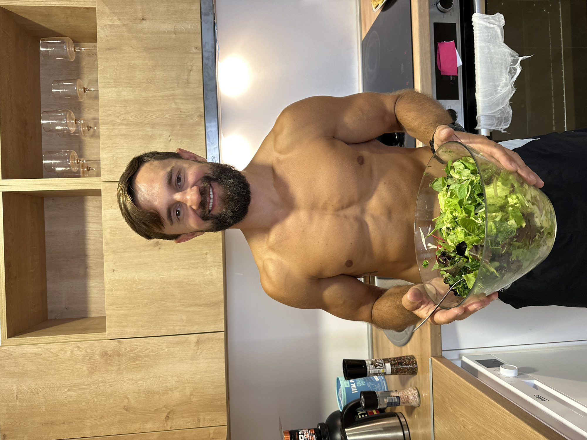 A shirtless man with a beard smiles at the camera, holding a large glass bowl of salad in a kitchen. The kitchen features light wood cabinets and white countertops. On the counter, there are various bottles and containers, including what appear to be spices and a nutritional supplement. Behind the man, a stovetop and oven are visible. The man is wearing dark shorts. The overall impression is one of health and domesticity, possibly in a residential setting.