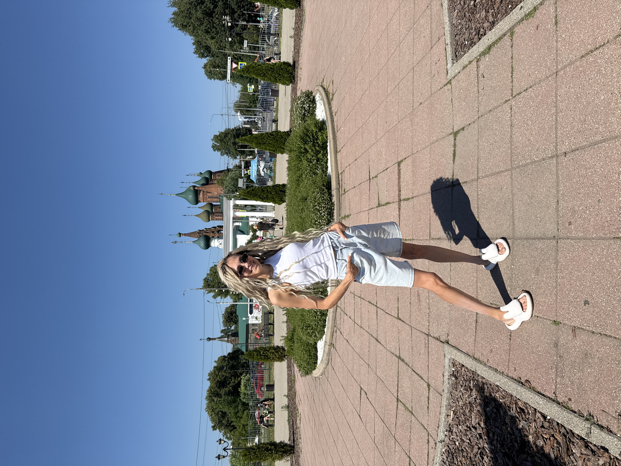 In the city of Zavolzhye, Russia, on a clear day, a woman with long, blonde dreadlocks and sunglasses stands in a paved outdoor area. She is wearing a white tank top and light blue denim shorts, with her hands in her pockets. White slide sandals are on her feet. Behind her, a circular garden bed with manicured bushes and white decorative stones is visible, bordered by manicured conical trees. In the background, a street with trees, a fence, streetlights, and traffic signs can be seen, along with a distinctive church with multiple green domes. The ground is paved with reddish-brown square tiles, and areas of dark mulch are present.