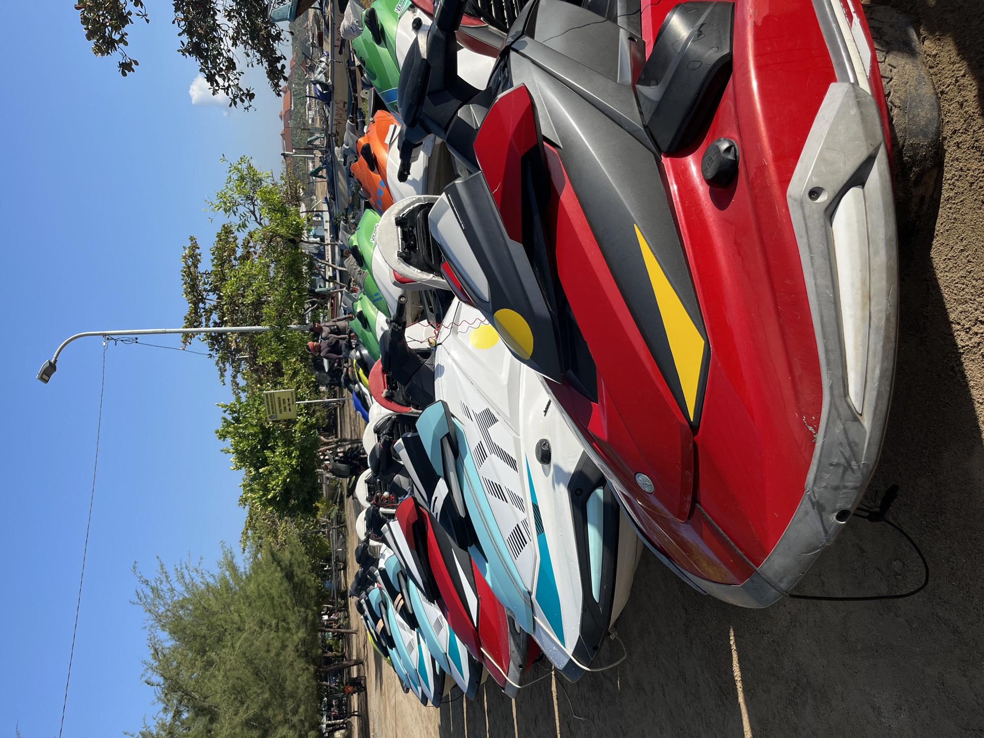 In Kuta Selatan, Indonesia, a scene unfolds on a sandy beach with a clear blue sky. A line of jet skis is parked, facing the viewer, on the sand. The jet skis are various colors, including red, white, blue, and green, with some featuring intricate patterns. In the background, trees and vegetation border the beach, and in the distance, buildings and more boats are visible. A few individuals are present, standing near the jet skis and in the background, appearing to be involved in recreational activities or operations. The overall environment suggests a popular tourist destination, likely a beach resort area.