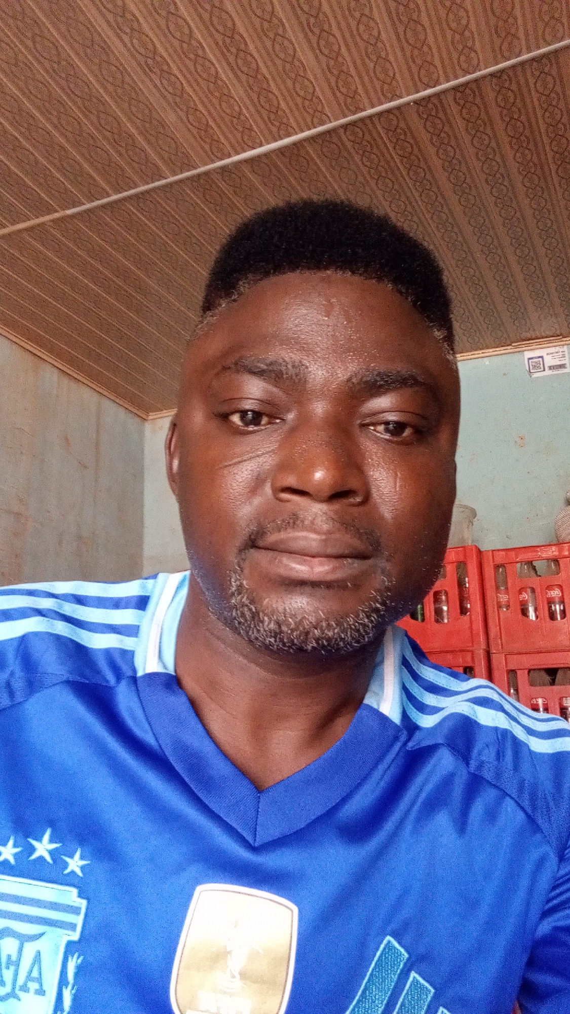 A close-up portrait shot features a man with dark skin, dark curly hair, and a salt-and-pepper beard, looking directly at the camera with a neutral expression. His face appears slightly shiny. Distinct scars are visible on his face, particularly on his left cheek (viewer's right).
He is wearing a bright blue short-sleeved collared shirt with a "V" neckline and light blue stripes across the shoulders. The shirt displays several emblems: on the left side of his chest (viewer's left), there is a partial logo that resembles the Argentine Football Association (AFA) crest, accompanied by three stars. On the right side of his chest, a prominent gold badge reads "FIFA World Champions."
The indoor background includes a brown ceiling with a repetitive patterned design. Behind the man, a light-colored wall is visible on the left. To his right, a stack of red plastic crates is present, some of which contain bottles, and partially visible text identifies them as "Coca-Cola" crates. A small sign with a QR code and some unreadable text is also visible on the wall in the upper right background. The setting suggests a casual indoor environment, possibly a shop or storage area.