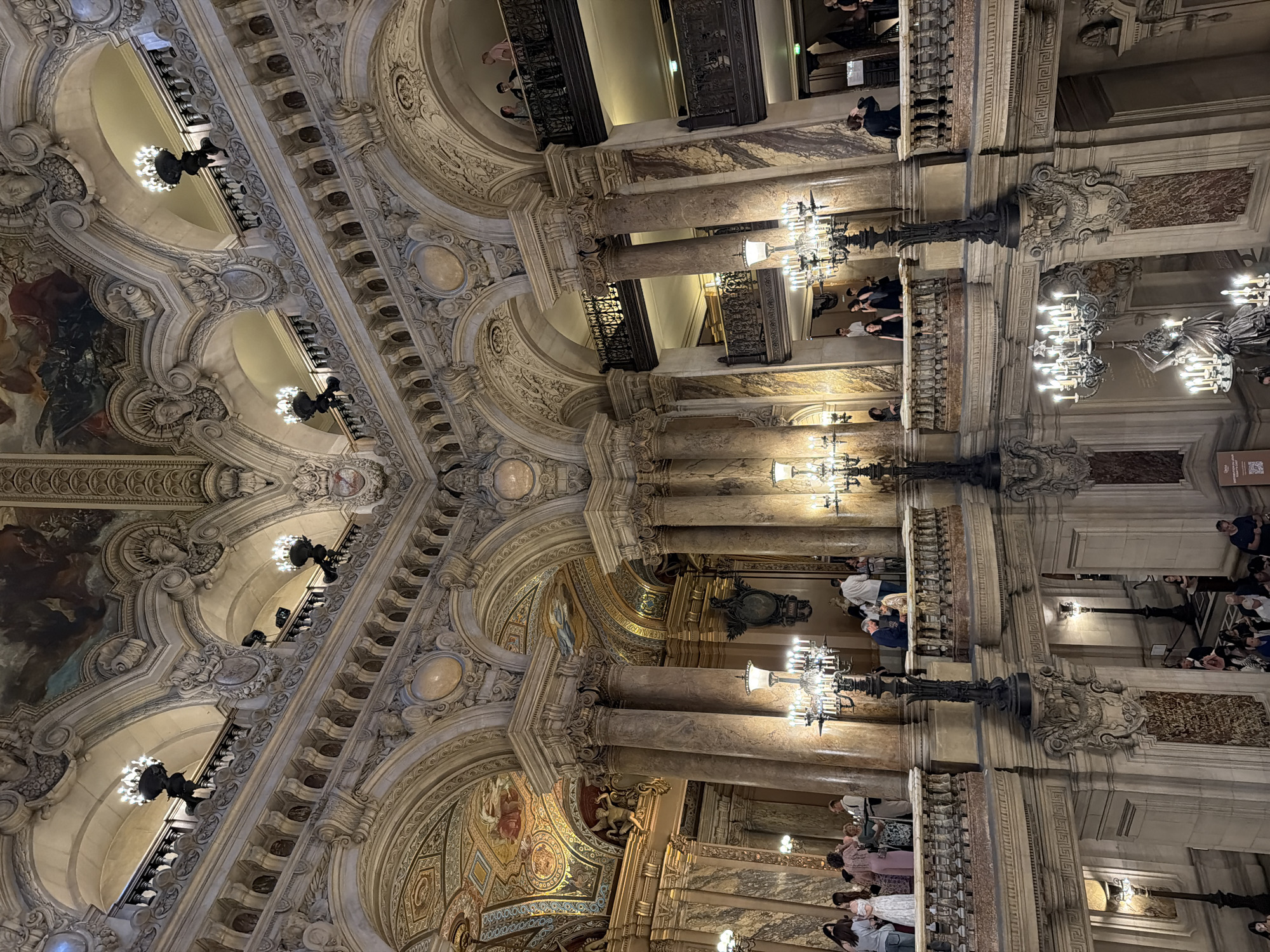 The image captures the opulent interior of the Palais Garnier, the Opera National de Paris, located in Paris, France. The ornate architecture showcases elaborate moldings, frescoes, and decorative elements characteristic of the Beaux-Arts style. Groups of people are visible on various levels, including balconies and staircases, observing the surroundings. Chandeliers provide illumination, accentuating the grandeur of the space. Marble columns and intricate detailing further enhance the overall impression of wealth and sophistication.