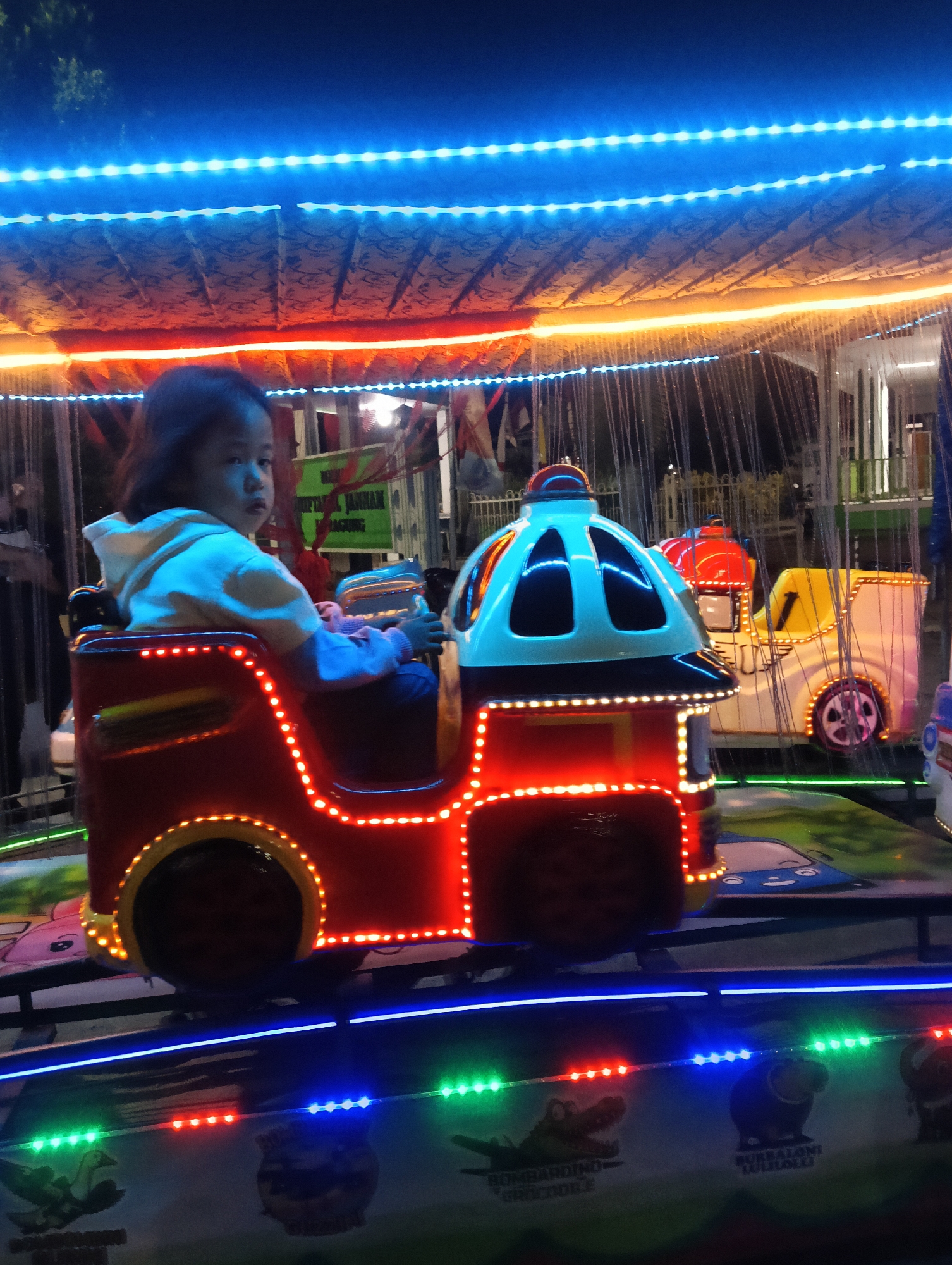 The image depicts a young child riding a miniature train at an amusement park in Luragunglandeuh, Indonesia. The child, dressed in a light-colored hoodie, is seated in the front car of the train, which is adorned with bright red and yellow lights. The train appears to be part of a carousel-like ride, with other similar cars visible in the background. The scene is illuminated by a profusion of colorful LED lights, creating a vibrant and festive atmosphere. The sky is dark, indicating it is nighttime. In the background, there are glimpses of other amusement park structures and fences. The ground beneath the train track is decorated with images of cartoon animals and text, suggesting a playful theme. The overall impression is one of a child enjoying a ride at a local fair.