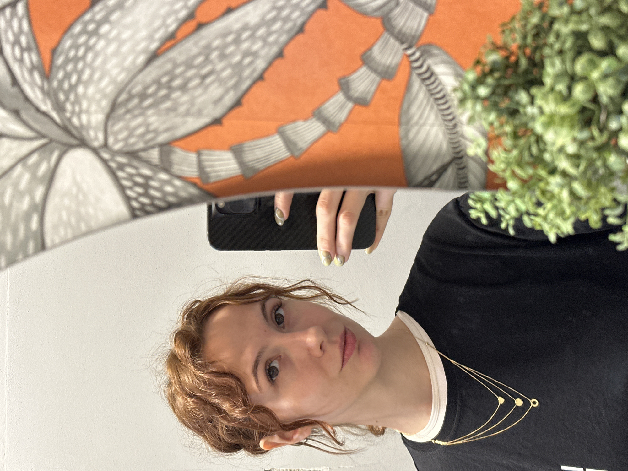 A mirror selfie taken indoors depicts a young woman with shoulder-length, curly reddish-brown hair and fair skin, looking directly at the viewer with a neutral expression. She is wearing a black t-shirt over a white top and a delicate, layered gold-colored necklace adorned with small circular and heart-shaped pendants. Her right hand, holding a black smartphone with a textured case, is visible above her head. Her fingernails feature a light, possibly yellow or green, nail polish with a subtle pattern.
The background directly behind the woman is a plain, light-colored wall. Above her, reflected in the mirror, is a vibrant orange surface featuring large, stylized gray and white botanical or abstract patterns, suggesting artwork or fabric. In the upper right corner of the reflection, a blurred artificial or real plant with small green leaves and reddish-brown accents is visible. The scene is brightly lit. The image is oriented sideways, with the woman's head to the left and her body extending to the right. No visible text is present in the image.