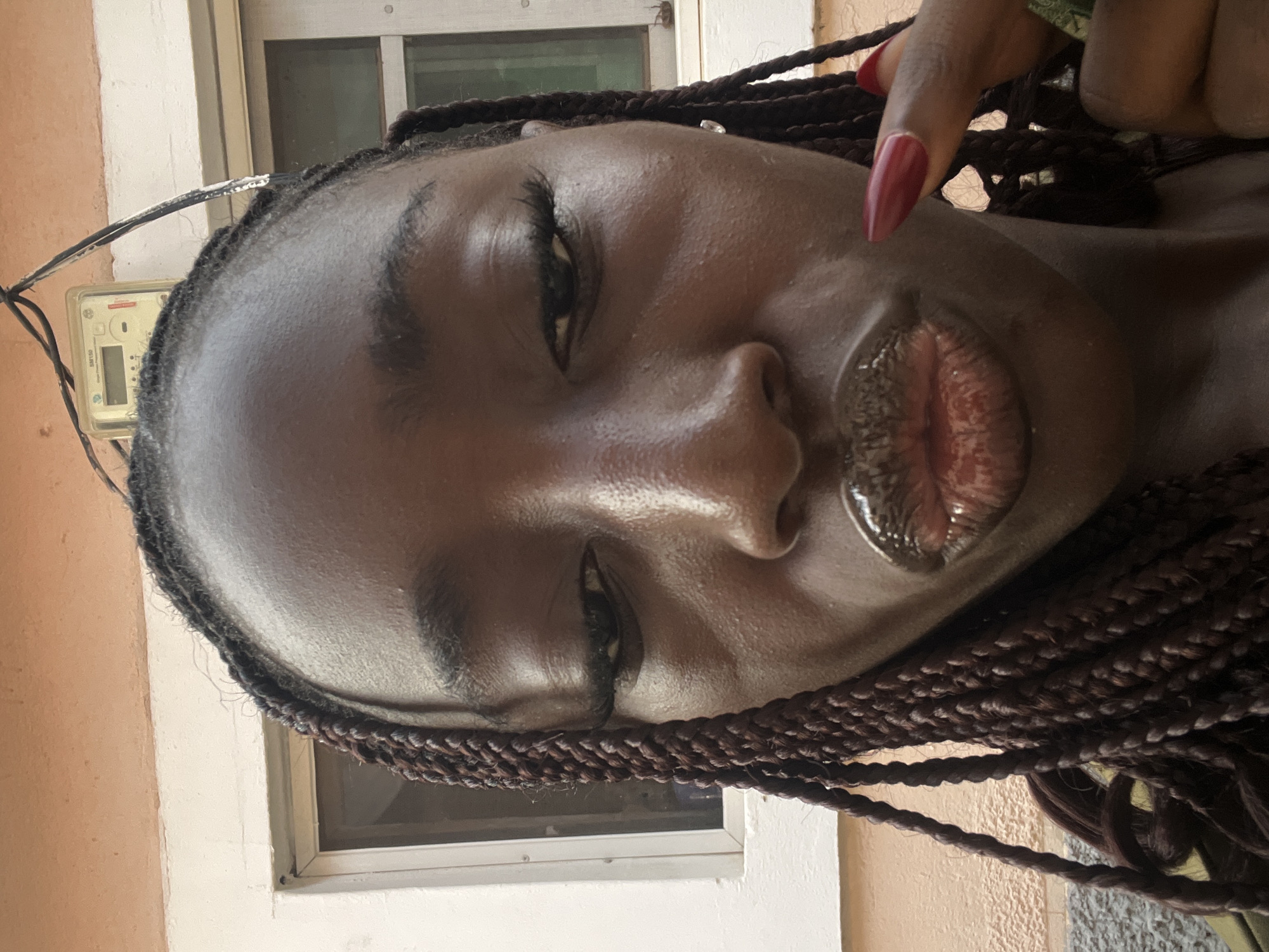 A close-up, slightly rotated, medium shot depicts a dark-skinned woman with glossy lips and braided hair. A red nail is visible in the upper right. Behind the woman is a light-colored wall with a window and an electrical meter. Given the location, the image was likely taken in Abuja, Nigeria.