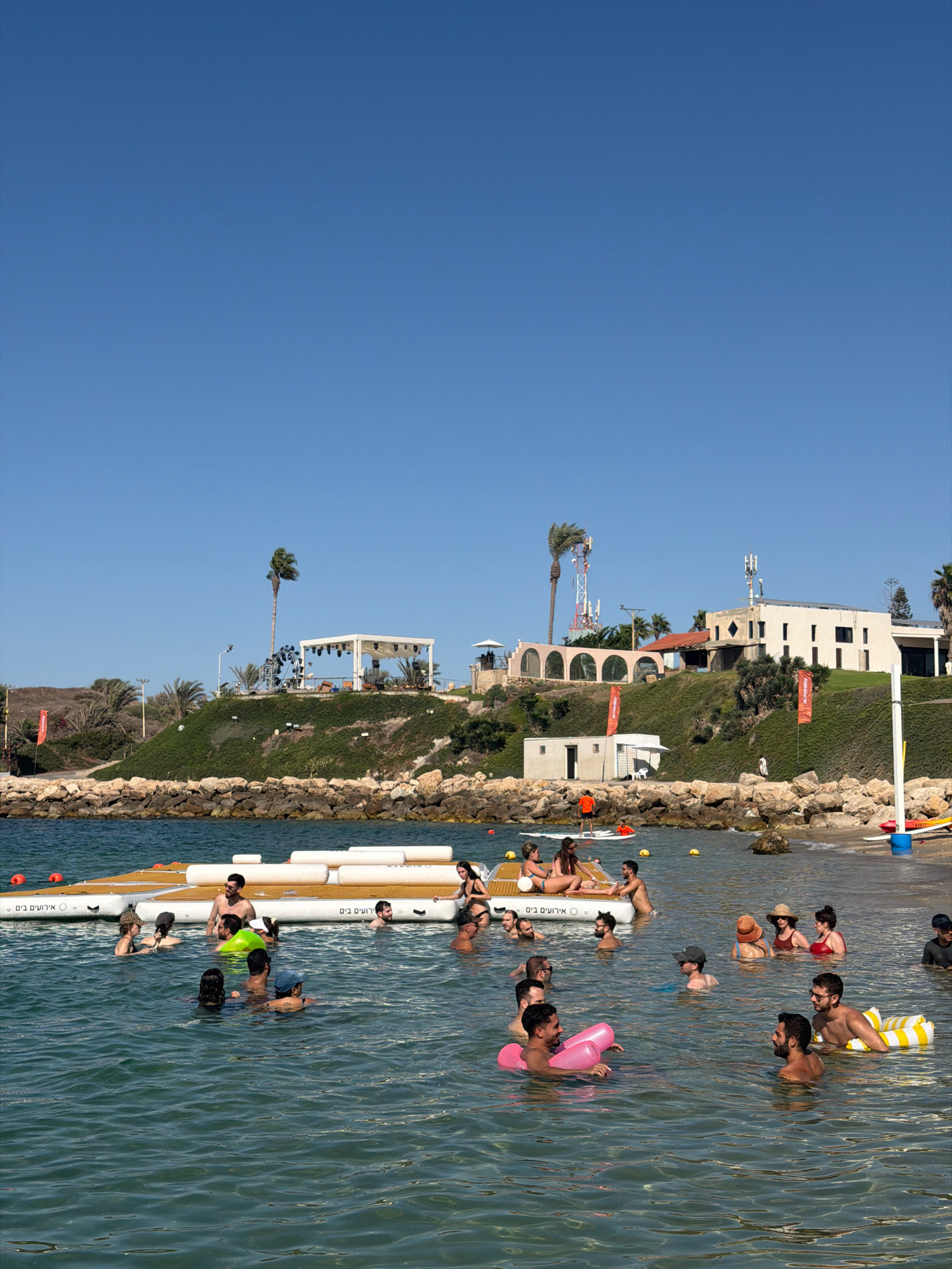 This is a medium-shot, eye-level, daylight, outdoor photograph taken at a beach in Hof HaCarmel Regional Council, Israel. The foreground features clear, turquoise water where several people are swimming and interacting. In the midground, there is a large floating platform with people sitting and standing on it. Behind the platform, there is a rocky shoreline with green vegetation and buildings perched on a hill. The buildings appear to be hotels or resorts, and there are palm trees visible. The sky is clear and blue.
Specifically, in the water, a group of individuals are enjoying a swim. Some are standing, others are submerged up to their torsos. One person is on a stand-up paddleboard, and another is holding a pink inflatable ring. Two men in the lower right foreground are facing each other, one with a yellow and white striped inflatable ring.
On the floating platform, several individuals are seated. A woman in a bikini is lying down, and others are socializing.
On the shore, a few structures are visible. There is a white, open-sided structure with a white canopy on the hill, possibly a stage or event area, with lighting equipment. Further to the right, a white building with multiple windows is situated on the hill. Two red flags with white text are planted on the grassy slope. In the distance, several cell towers are prominent. The rocky beach is lined with stones.