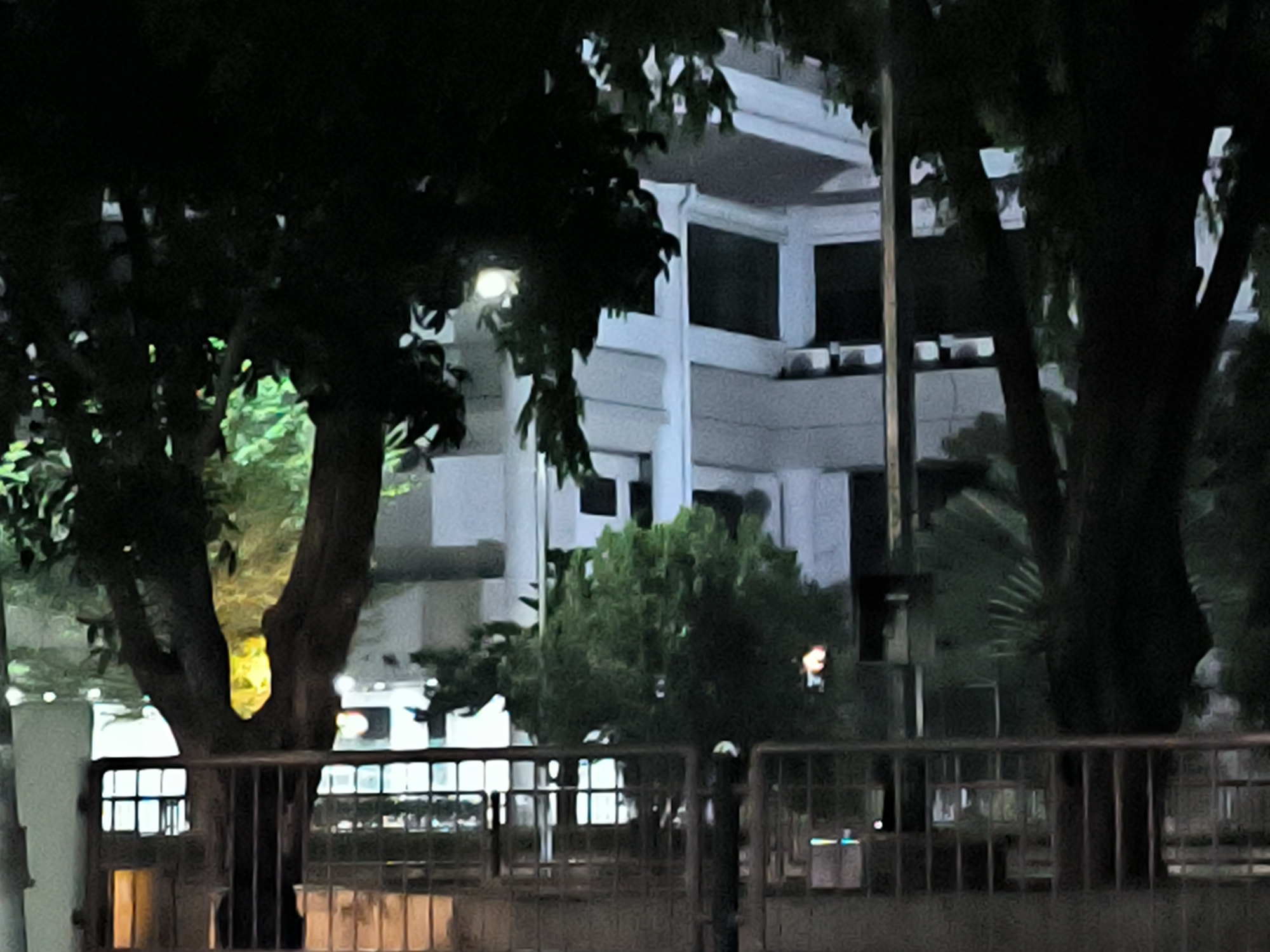 The image shows a building viewed through trees and a metal fence. The building is several stories tall with a white facade and dark windows. Trees obscure portions of the building. A metal fence runs along the bottom of the frame. The image appears to be taken at night or in low light conditions. The location is Lhokseumawe, Indonesia.