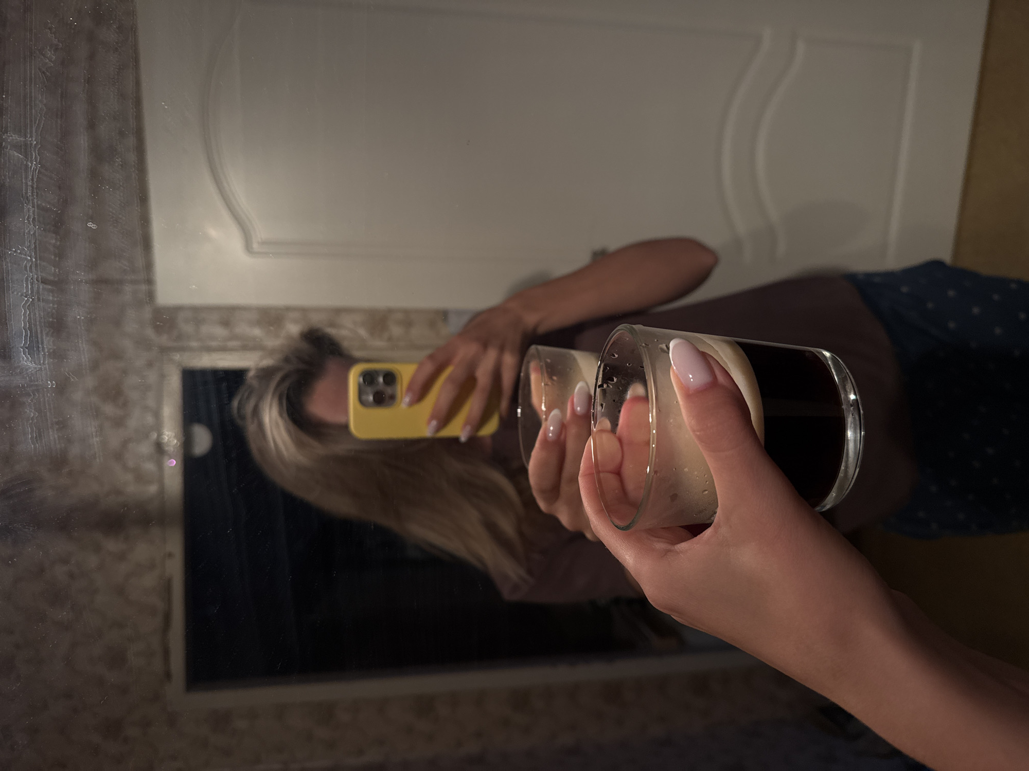 The image shows a mirror selfie taken indoors, reflecting a person holding a smartphone and a glass. The person, whose face is largely obscured, has light-colored hair and manicured nails with light pink/white tips. In their left hand (as seen in the reflection), they hold a smartphone with a distinctive yellow case and a triple camera lens setup, appearing to be the device used to take the photo. In their right hand, they hold a clear glass containing a dark liquid topped with a thick layer of light-colored foam, possibly a stout beer or a coffee drink.
The reflection reveals part of the person's clothing: a dark top and a dark blue garment with white polka dots. The mirror itself shows some smudges and streaks, particularly on the left side. Behind the person's reflection, a light-colored wall or paneling with decorative, curved molding is visible. The overall lighting is dim, suggesting an indoor evening or nighttime setting. No specific location cues are present beyond an indoor domestic environment. There is no visible text in the image.