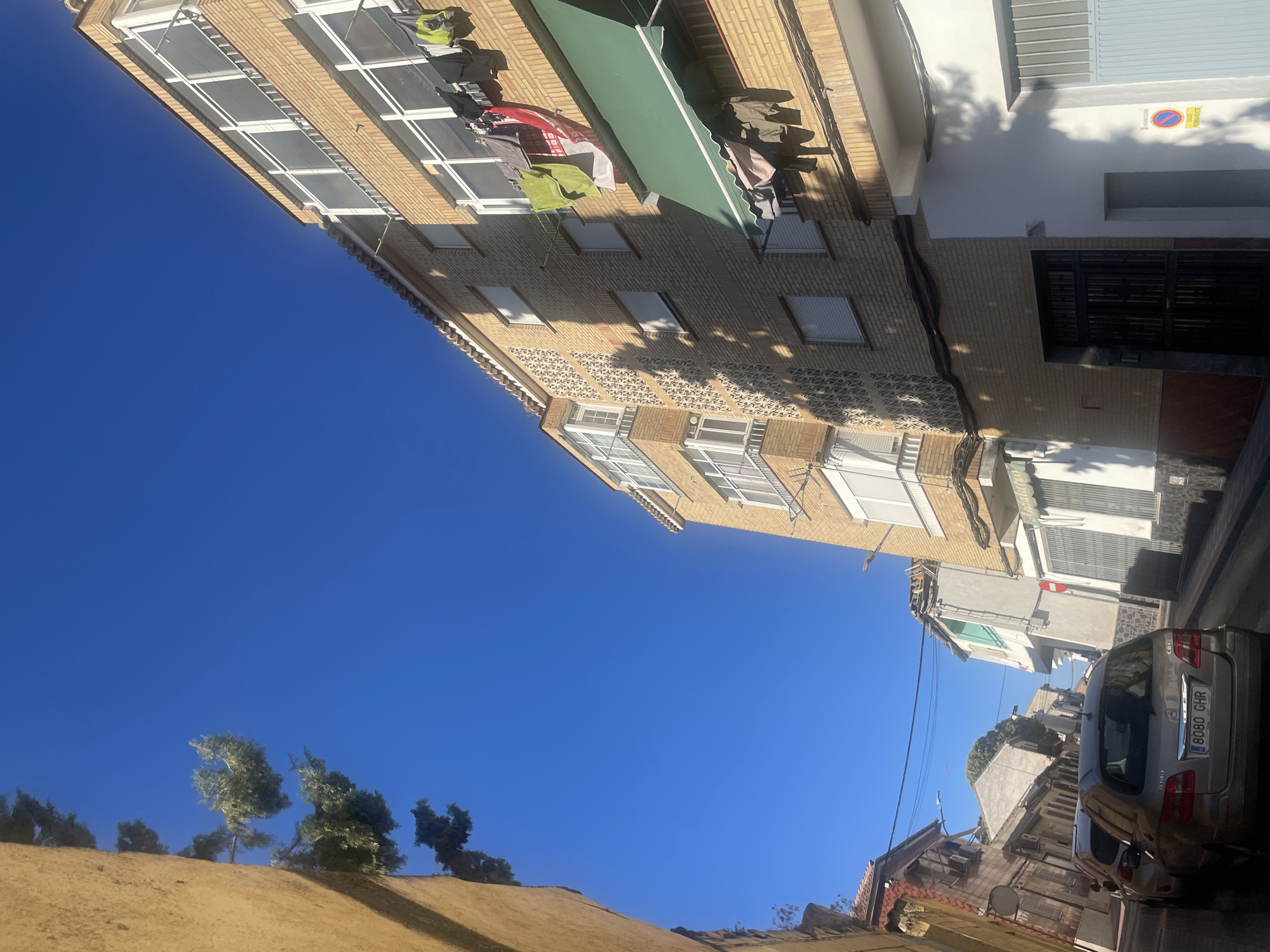 The image is a low-angle shot looking up at a narrow street in Atarfe, Spain. The sky is clear and blue. On the right side of the street, a multi-story brick building dominates the view. Clothes are hanging from lines on the balconies of this building, including a green towel, a red and white striped sheet, and several pieces of clothing in shades of grey and black. A green awning extends from one of the balconies. Below this building, a white wall with a dark metal gate is visible. On the left side of the street, another building made of rough-textured, light-colored material forms a wall that extends upwards. Trees with green foliage hang into the frame from above this wall. Further down the street, other buildings with various architectural styles and colors can be seen, some with windows and balconies. Two cars are parked on the street, with the rear of a silver Mercedes-Benz station wagon prominently in the foreground, showing a license plate that reads "8080 GHR". Electrical wires crisscross above the street. The overall impression is of a residential street in a Spanish town on a bright, sunny day.