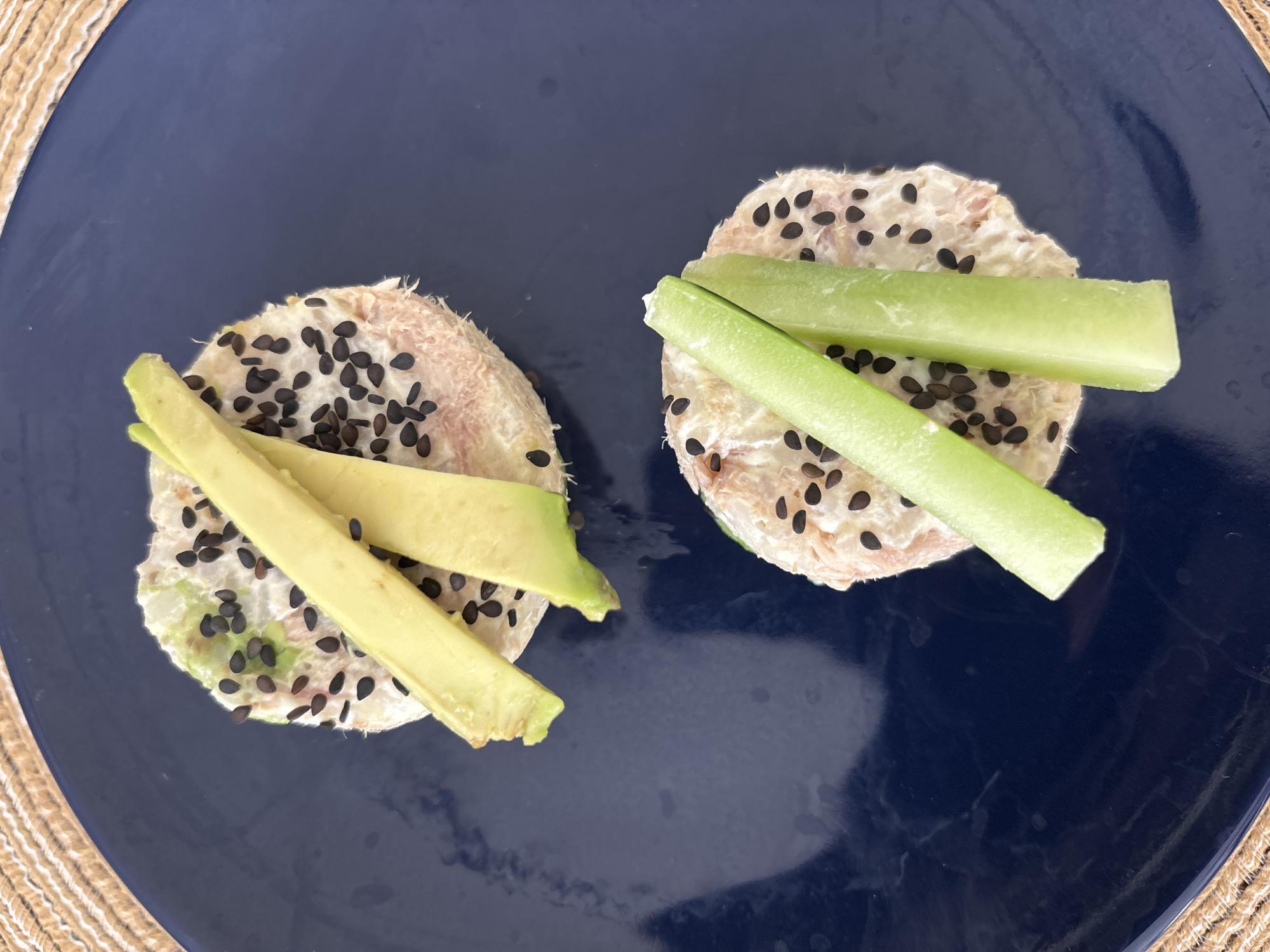Two small, circular stacks of food are arranged on a dark blue plate. Each stack is topped with black sesame seeds. One stack has two strips of avocado on top, while the other has two strips of a green vegetable, possibly cucumber or celery. The plate rests on a tan woven surface. The location is not evident from the image.
