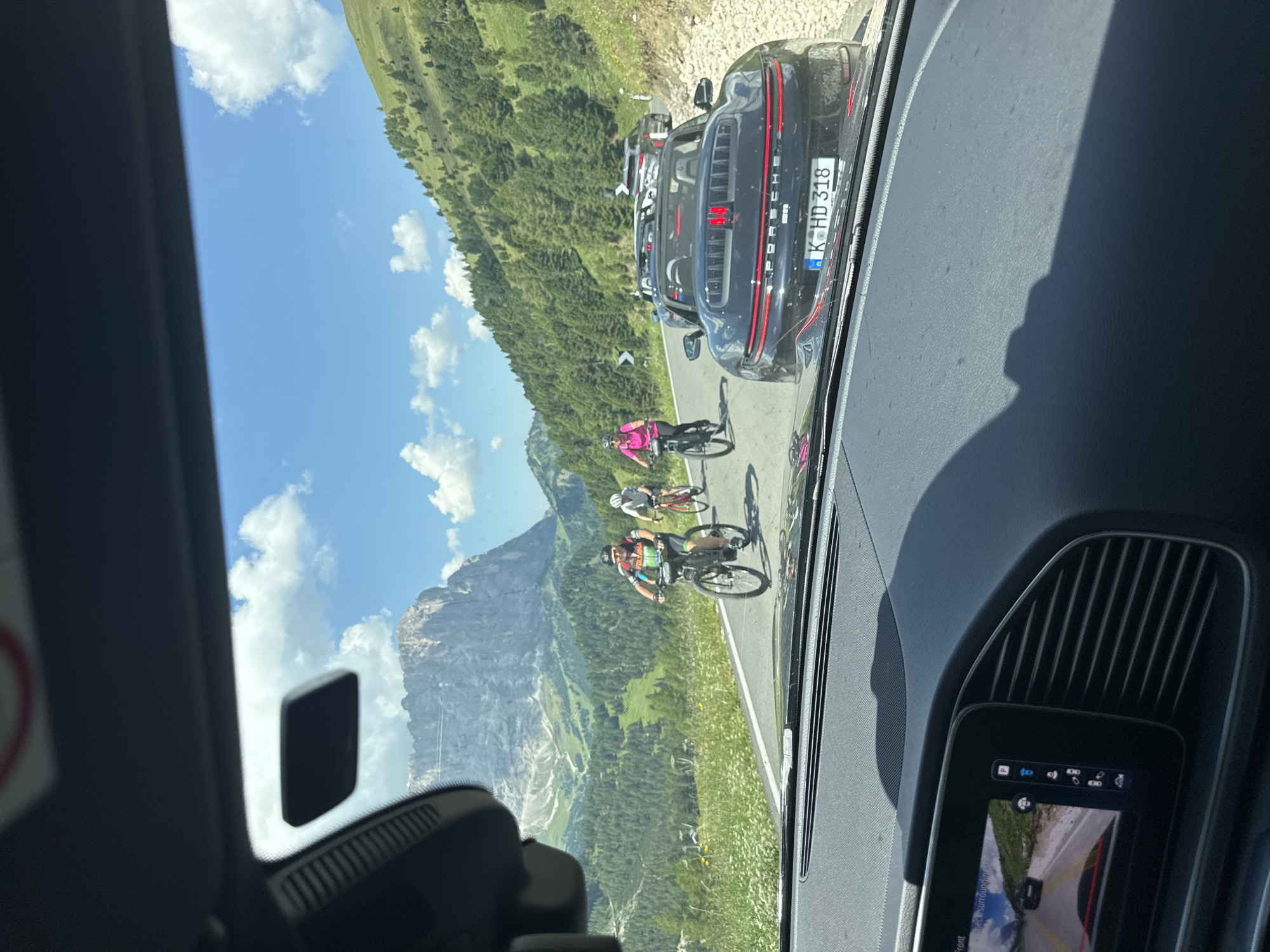 The image, taken from the interior of a vehicle, captures a scenic mountain road in Sëlva - Wolkenstein - Selva di Val Gardena, Italy. The road is populated with several cyclists and cars. In the foreground, a black convertible Porsche with a license plate reading "K-HD 318" is visible. Behind it, other cars are seen. Several cyclists are riding on the road. One cyclist, wearing a bright pink top and helmet, is on a black bicycle. Another cyclist, in a black and colorful jersey and helmet, is on a bicycle. A third cyclist, wearing a white helmet, is on a red bicycle. The road winds through a lush green mountainous landscape with trees and white wildflowers. In the background, a towering, rugged mountain dominates the skyline under a blue sky with scattered white clouds. The interior of the vehicle shows a dashboard with a screen displaying a rearview camera feed.