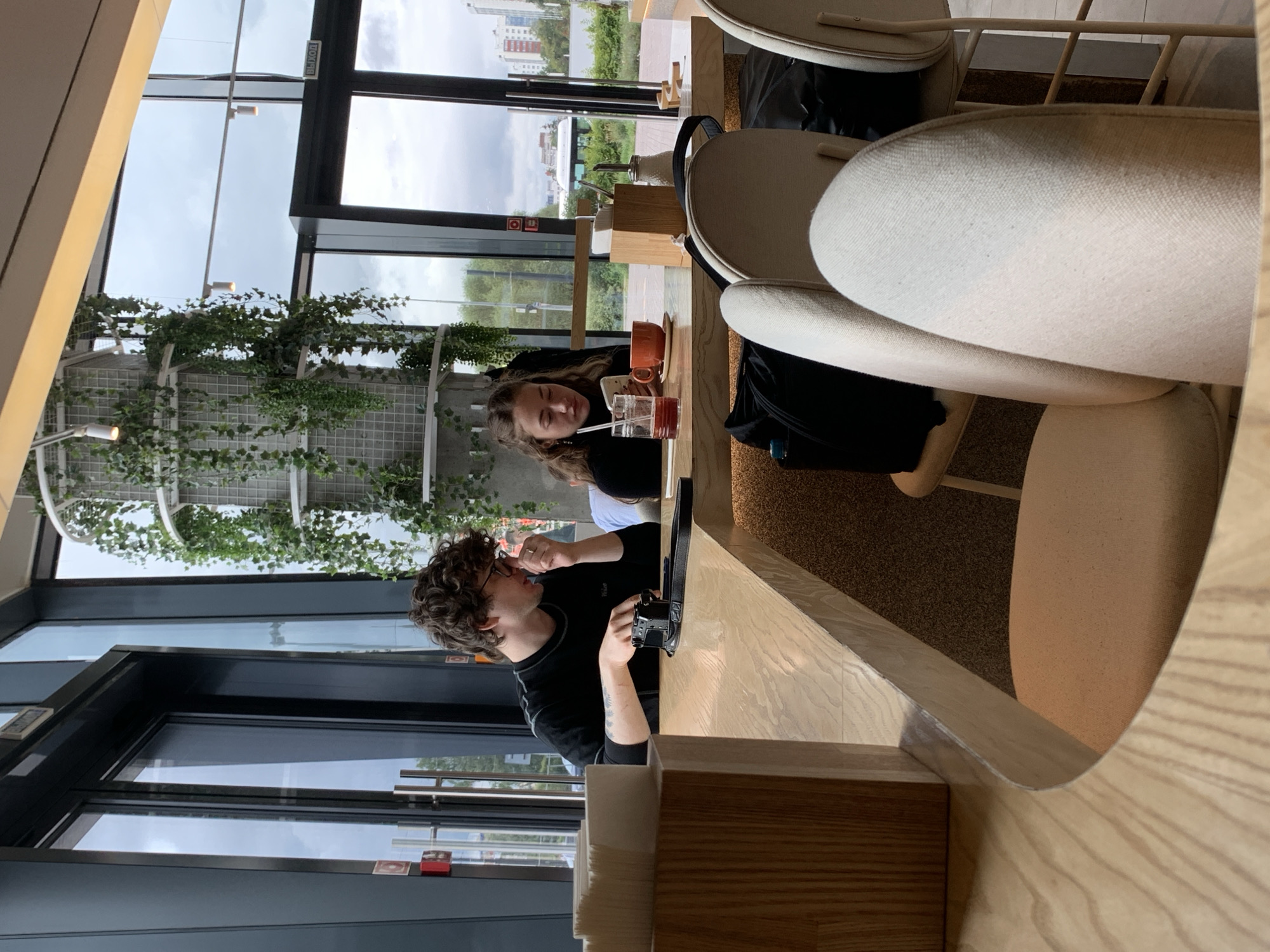 The image depicts an interior scene at a cafe, potentially located in the city of Perm, Russia, given the presence of the word "ВЫХОД" (Exit) on a sign visible through the glass doors. Two individuals, a male and a female, are seated at a light-colored wooden table. The male, with curly brown hair and glasses, is wearing a dark long-sleeved shirt and appears to be holding a small camera. The female, with long blonde hair, is wearing a dark top and is looking down at her phone, with a glass of red liquid and an orange cup on the table in front of her. Behind them, large windows offer a view of an outdoor area with greenery and a cloudy sky. The cafe interior features modern design elements, including hanging plants and light-colored chairs.