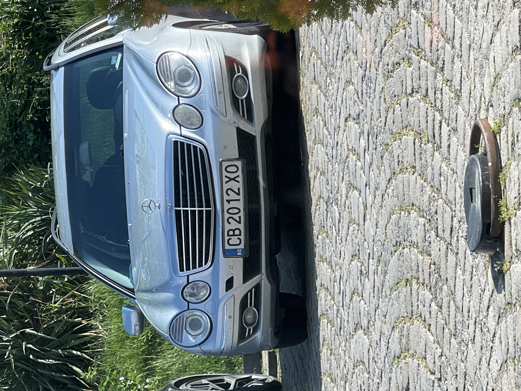The image shows the front of a silver Mercedes-Benz car parked on a stone-paved surface. The car's license plate reads "CB 2012 XO" and has the letters "BG" indicating it is registered in Bulgaria. The car is parked near a grassy area. To the right of the car is a dark-colored metal fixture that appears to be a bollard or a parking lock. The scene suggests an urban environment, possibly a street or parking area. The car's registration "CB" suggests the city of Sofia, Bulgaria.