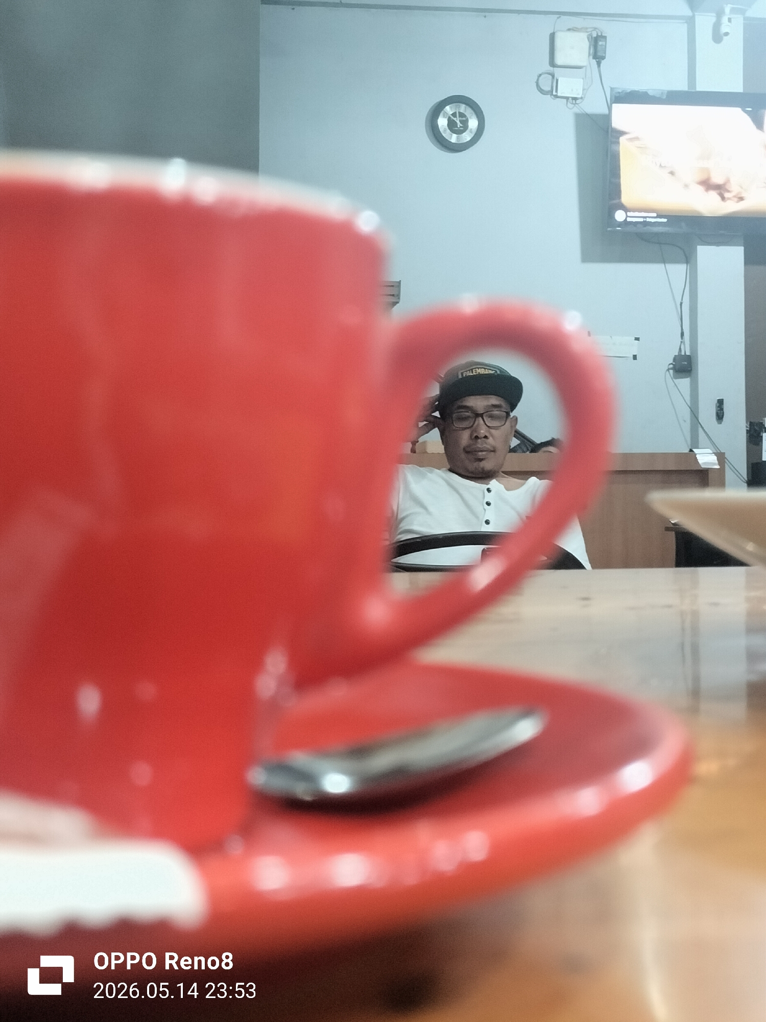 The image depicts a blurry foreground of a red coffee cup and saucer with a spoon, set on a wooden table. Through the handle of the cup, a man wearing a black "PALEMBANG" cap and glasses is visible, seated and looking forward. Behind him, a wall is visible with a clock, electrical box, and a television screen displaying a light-colored image. The image was taken using an OPPO Reno8 on May 14, 2026, at 23:53. There is no direct reference to Lhokseumawe, Indonesia, or any other specific location.