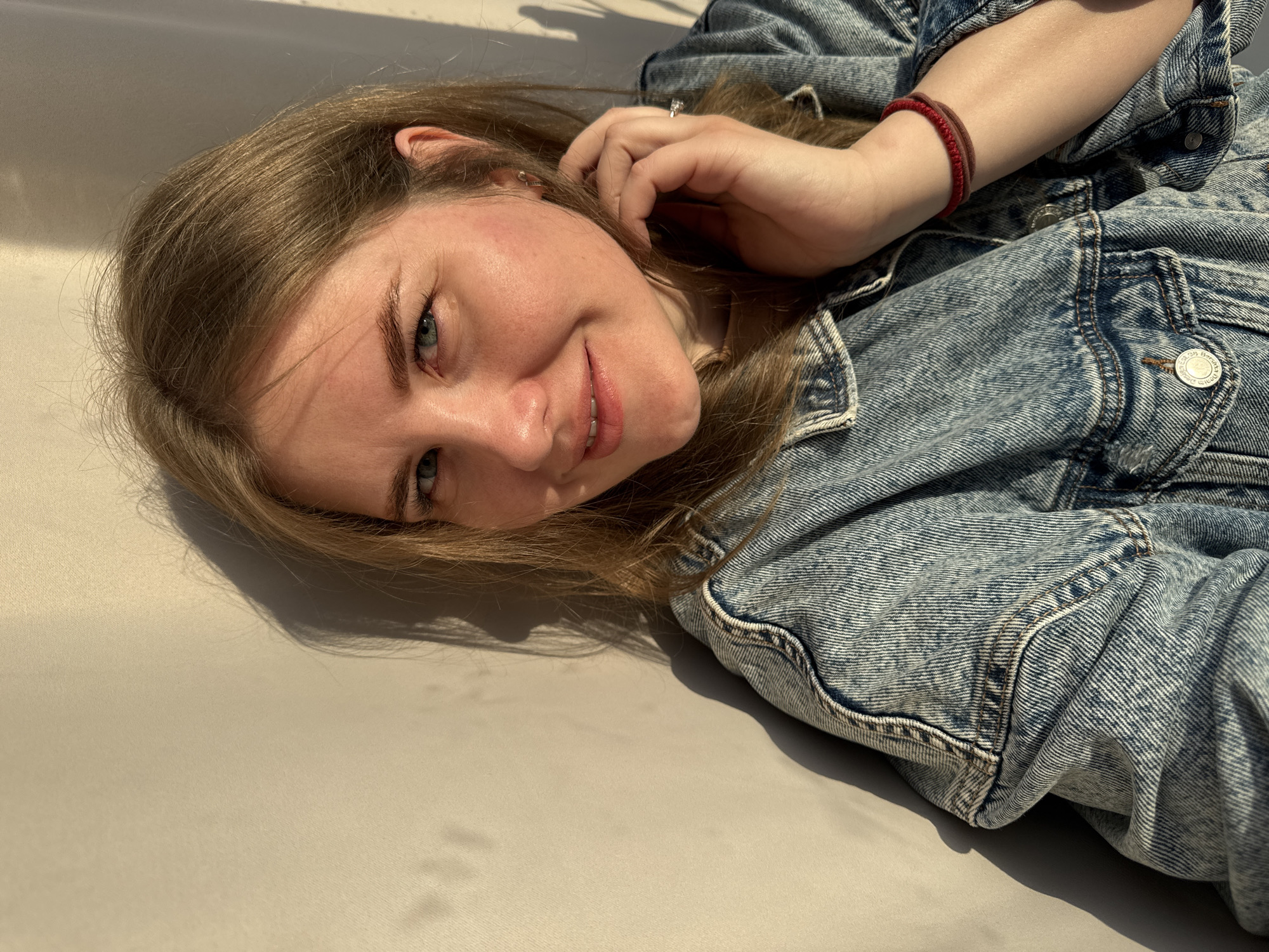 A fair-skinned woman with light brown hair lies on a flat, neutral-colored surface, possibly a bed or bench. She wears a blue denim jacket and a red bracelet on her right wrist. Her left hand is near her head, touching her hair. She has light eyes and is smiling slightly. The light source is from the side, casting shadows on the surface she is lying on. The location is not determinable from the image.