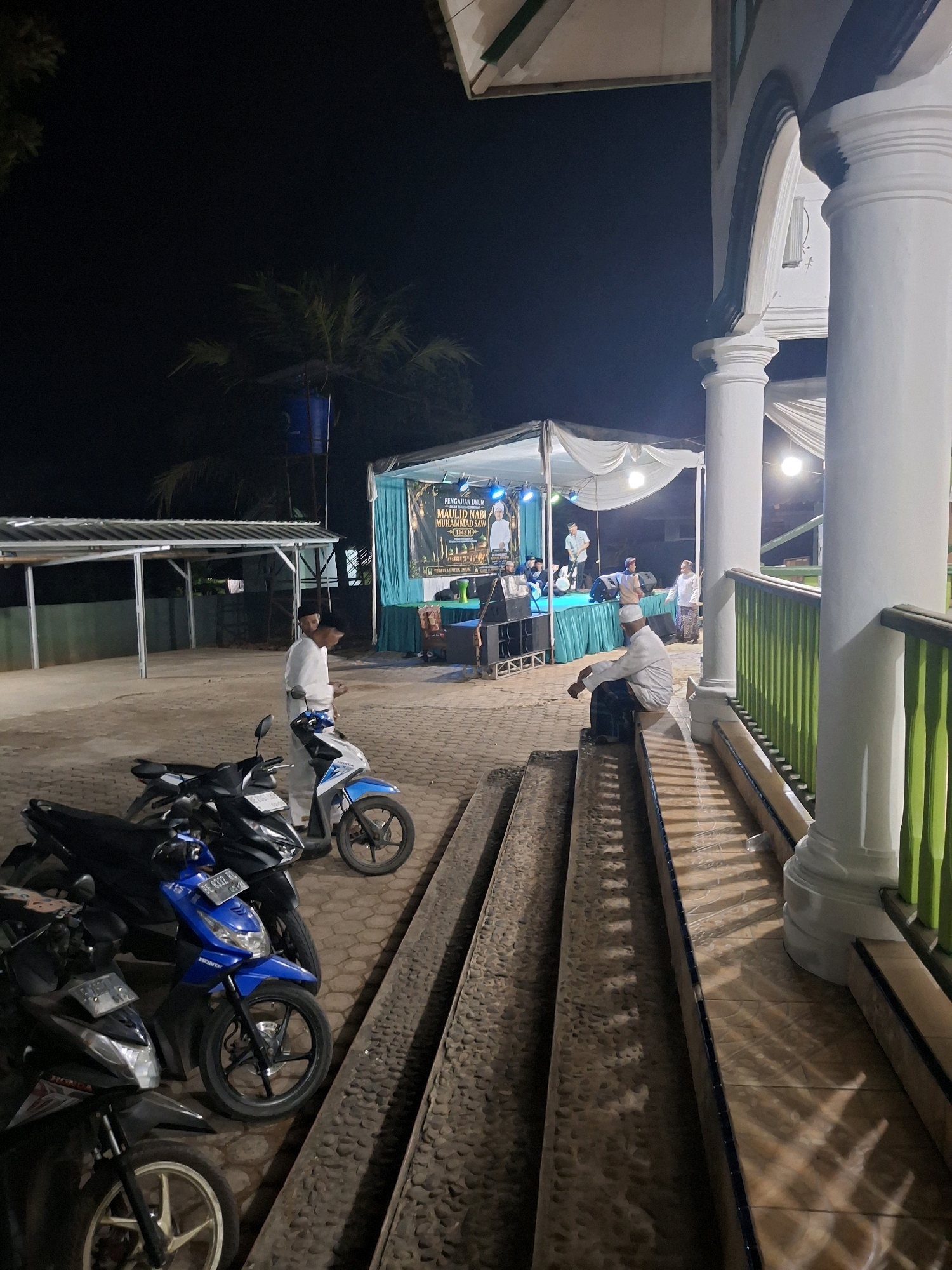 The image depicts an outdoor event at night, possibly a religious gathering, near a building with white pillars and a green railing. In the foreground, several motorcycles are parked on a cobblestone ground. To the left, there is a covered area with a metal roof. Further back, a stage is set up with a banner displaying text in Indonesian, and several people are visible on and around the stage. One person is sitting on steps in front of the building, facing away from the camera. The overall ambiance suggests a community event taking place in Yogyakarta, Indonesia.