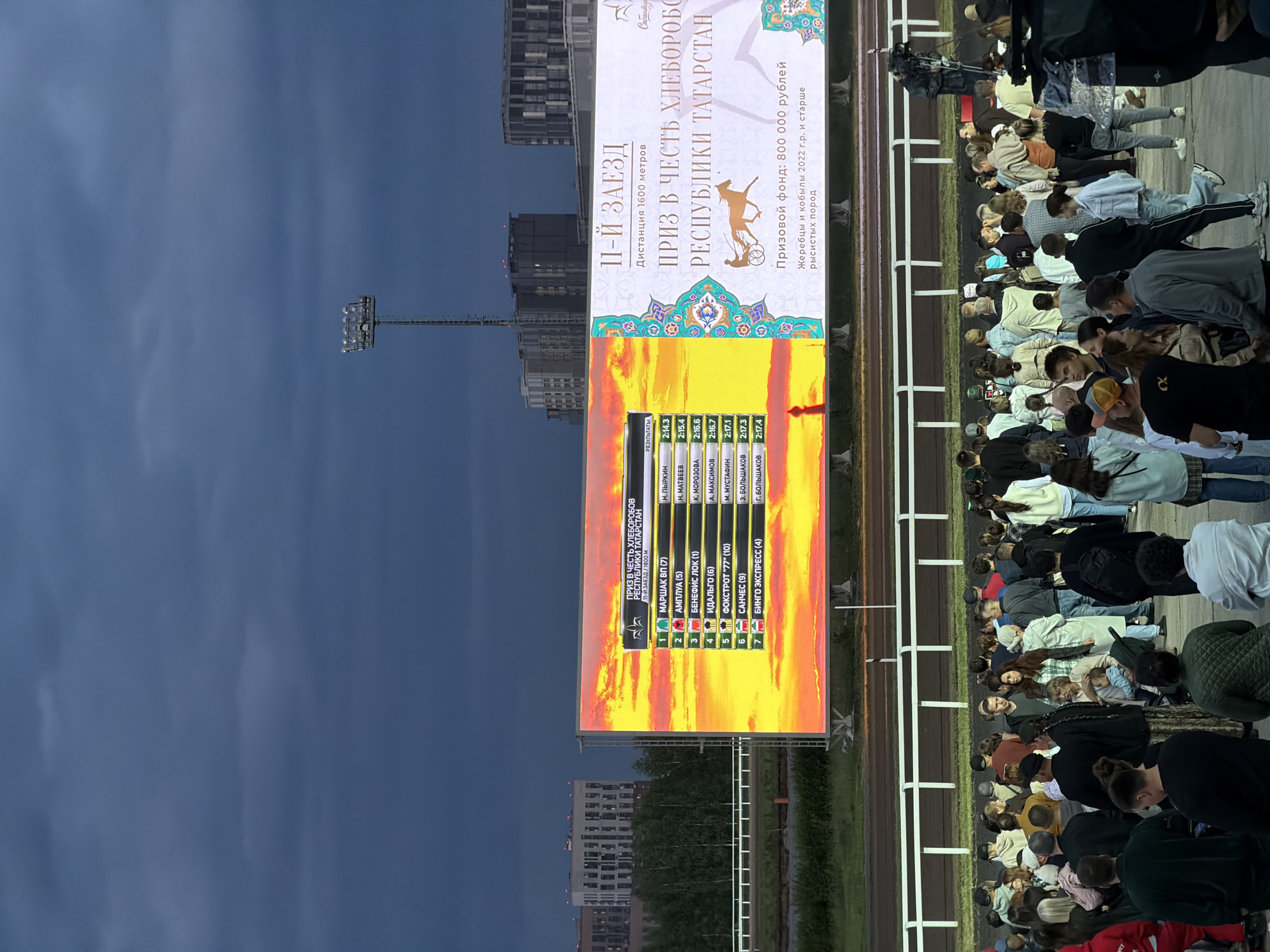The image shows a crowd of people gathered at a racetrack, looking at a large electronic billboard. The billboard displays results from a horse race, with horse names, jockeys, and finishing times listed. The race appears to be the 11th race, covering a distance of 1600 meters, and is part of a prize in honor of the Republic of Tatarstan's agricultural workers. The prize fund is 800,000 rubles. The weather appears overcast with a dramatic sky, suggesting it might be evening. The crowd is diverse, with individuals of various ages and backgrounds. Some are casually dressed, while others appear to be professional photographers or videographers, with cameras visible in the foreground. The racetrack itself is visible in the background, with white railings bordering the track. In the distance, modern buildings can be seen, indicating an urban or suburban setting.